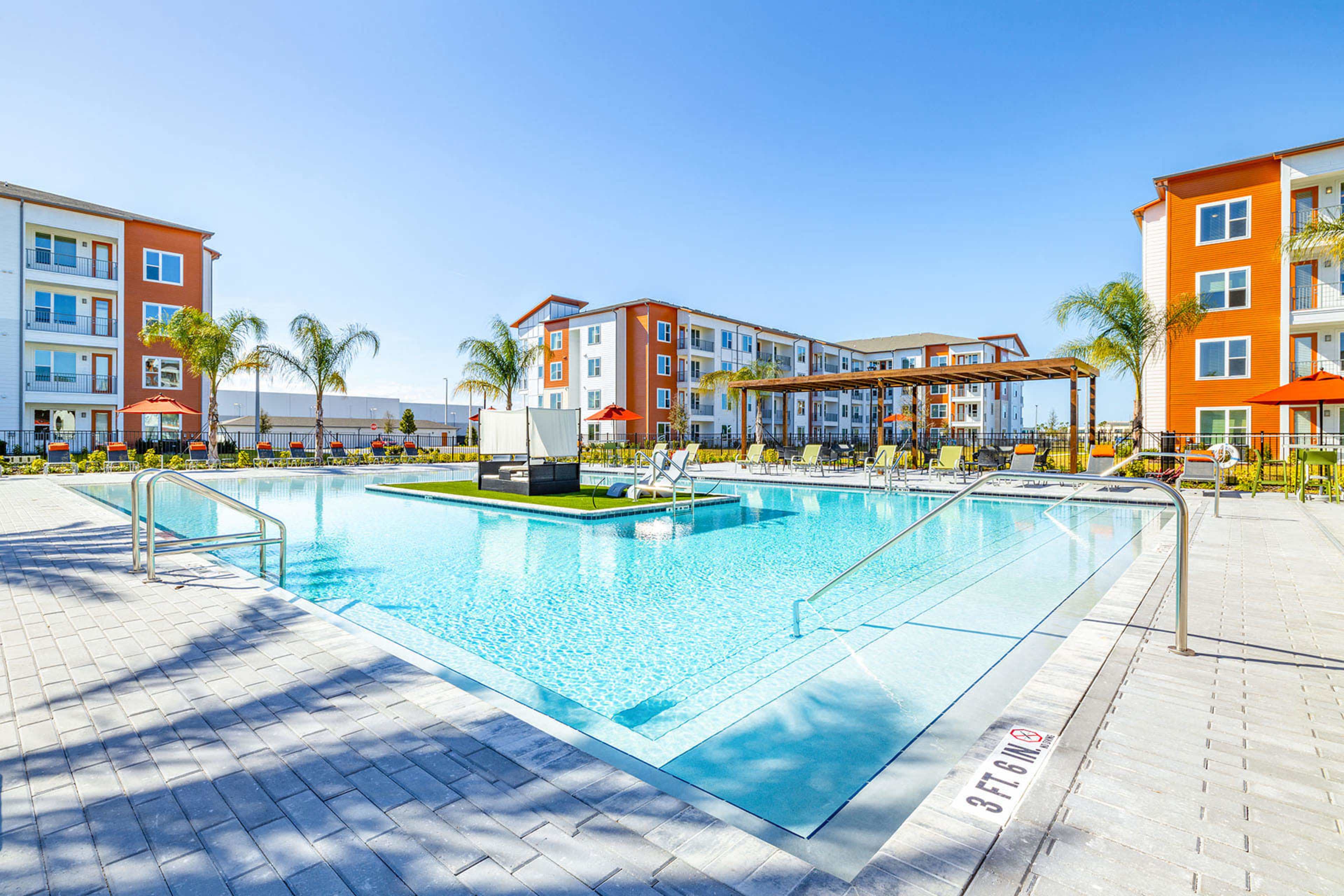 The image shows a modern apartment complex with a large swimming pool surrounded by palm trees and lounge areas.