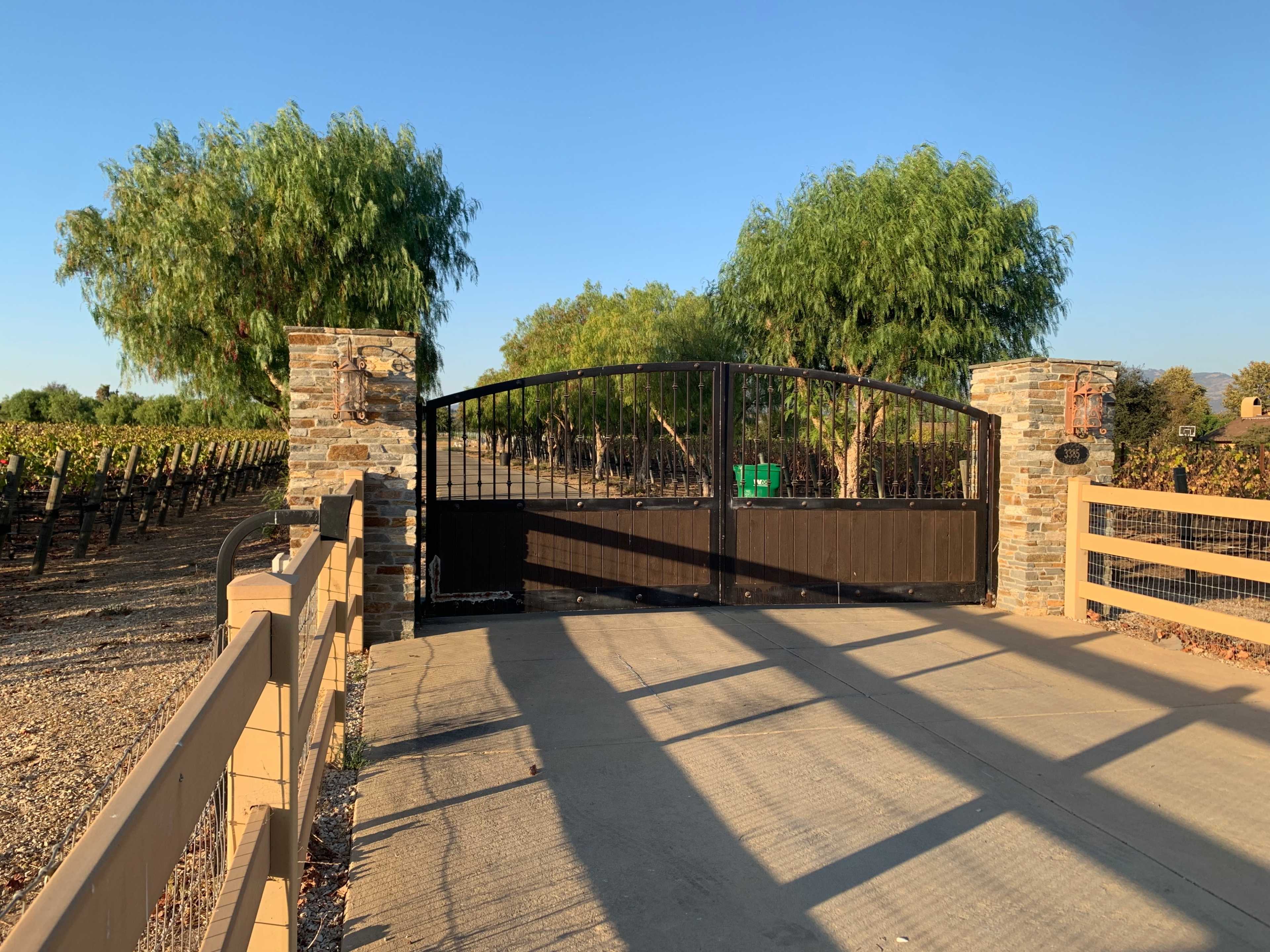 A metal gate stands at the entrance of a vineyard, flanked by stone pillars and lined with trees.