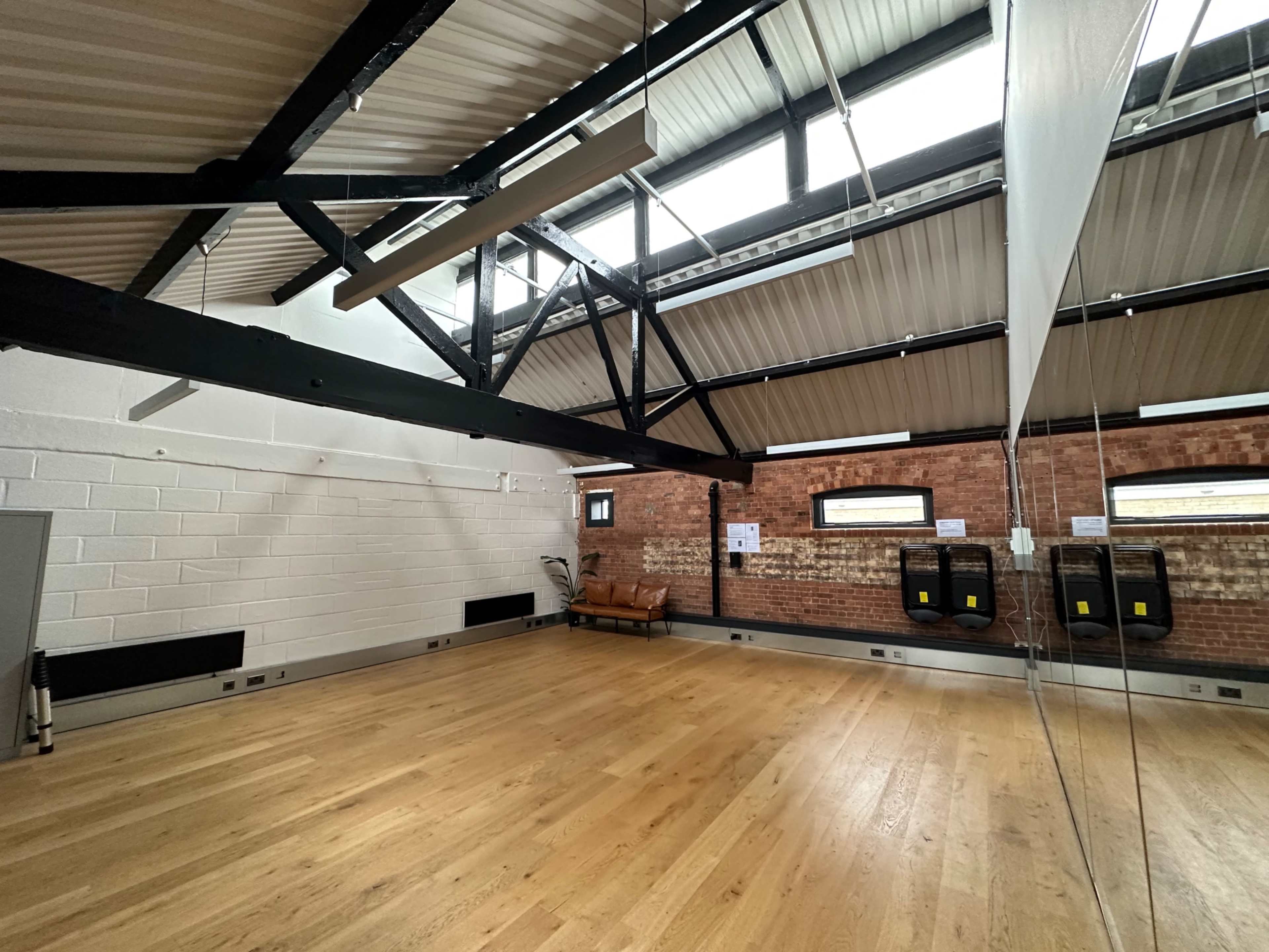 A spacious room with wooden flooring, exposed brick walls, and large windows under a metal beam ceiling.