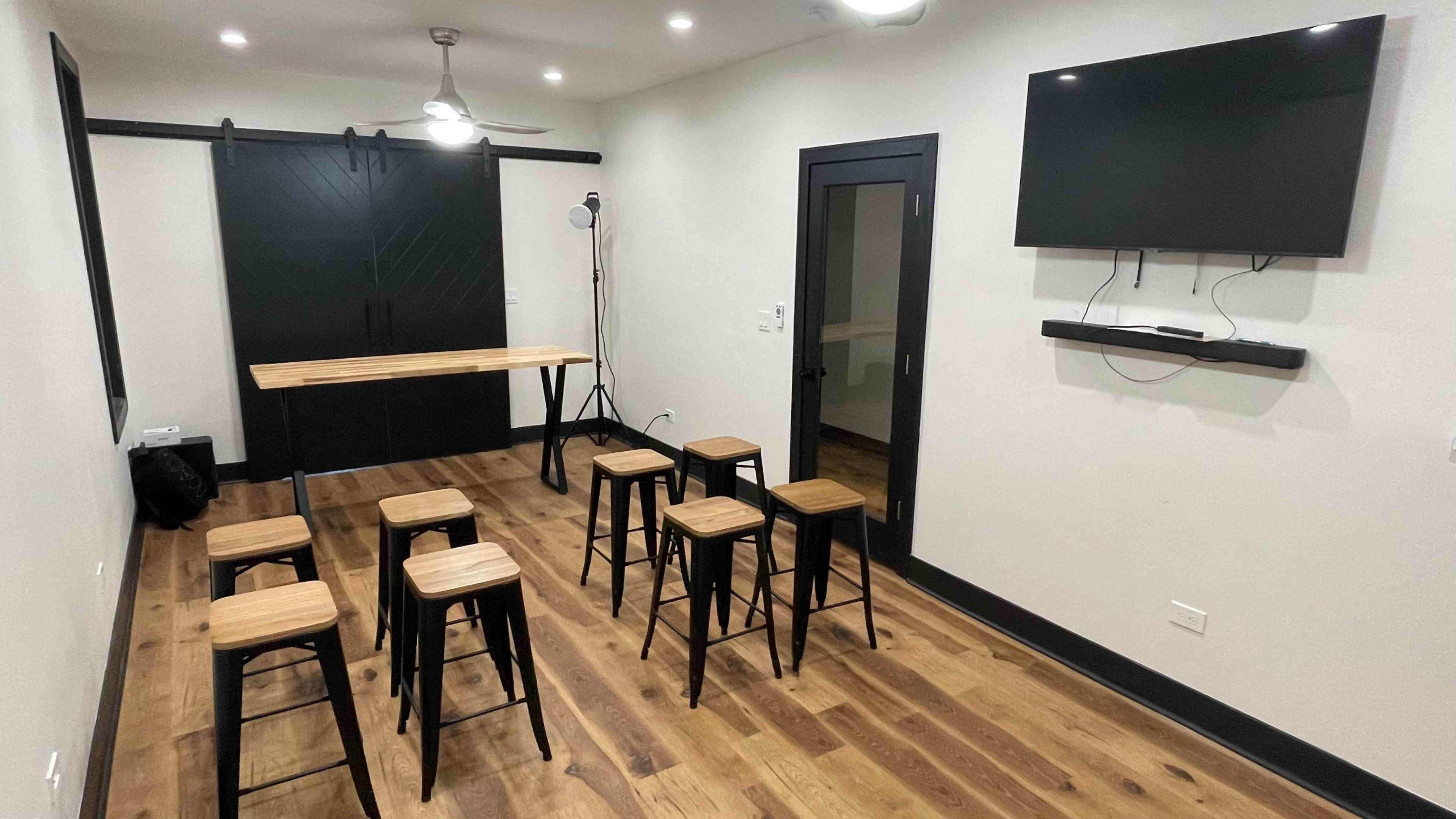 A modern meeting room features a long wooden table surrounded by eight black stools, with a wall-mounted TV and sliding barn door.