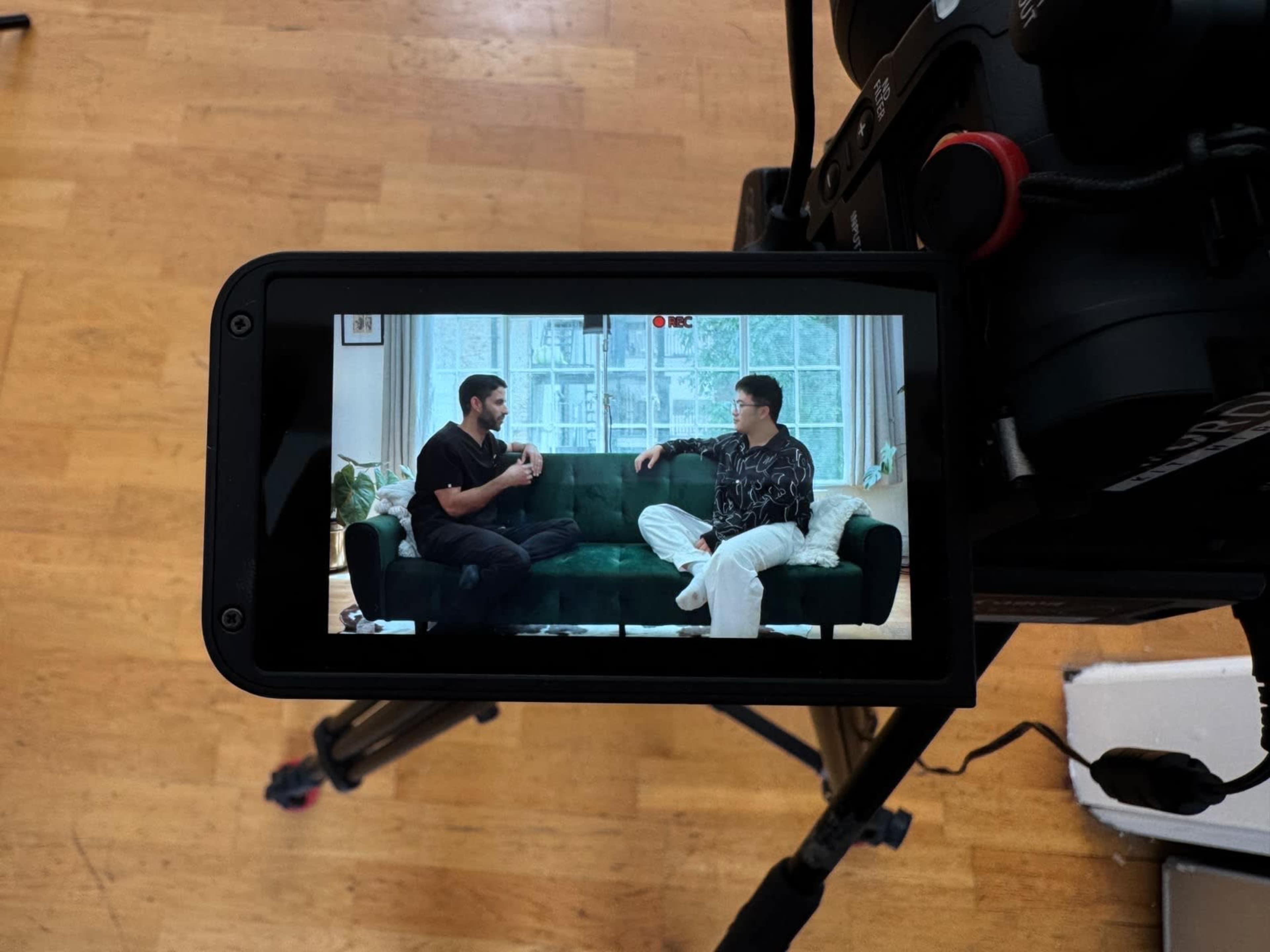 Two men are seated on a green couch in a well-lit room, engaged in conversation, while being filmed from above.