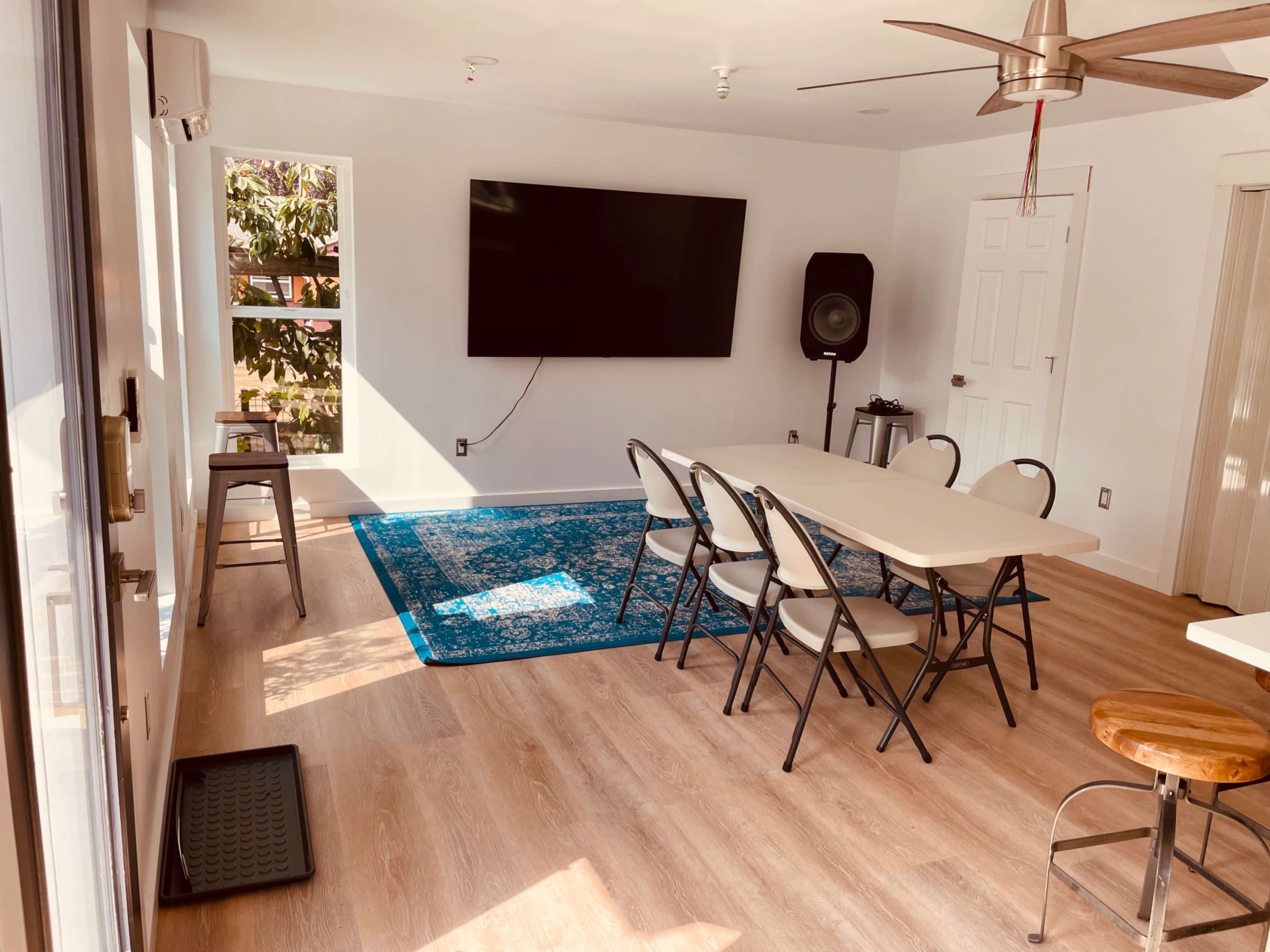 A spacious room features a large flat-screen TV on the wall, a long table surrounded by chairs, and a blue area rug on the wooden floor.