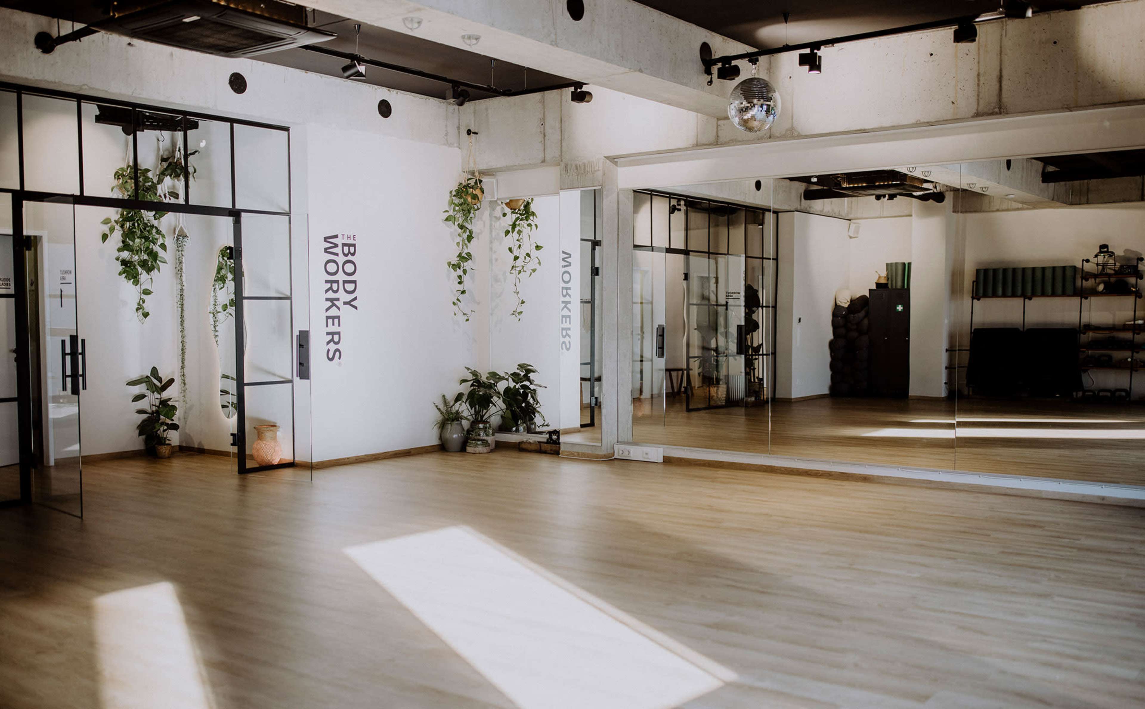 The image shows a spacious, modern studio with large mirrors, natural light, and plants, featuring the words "THE BODY WORKERS" on one wall.