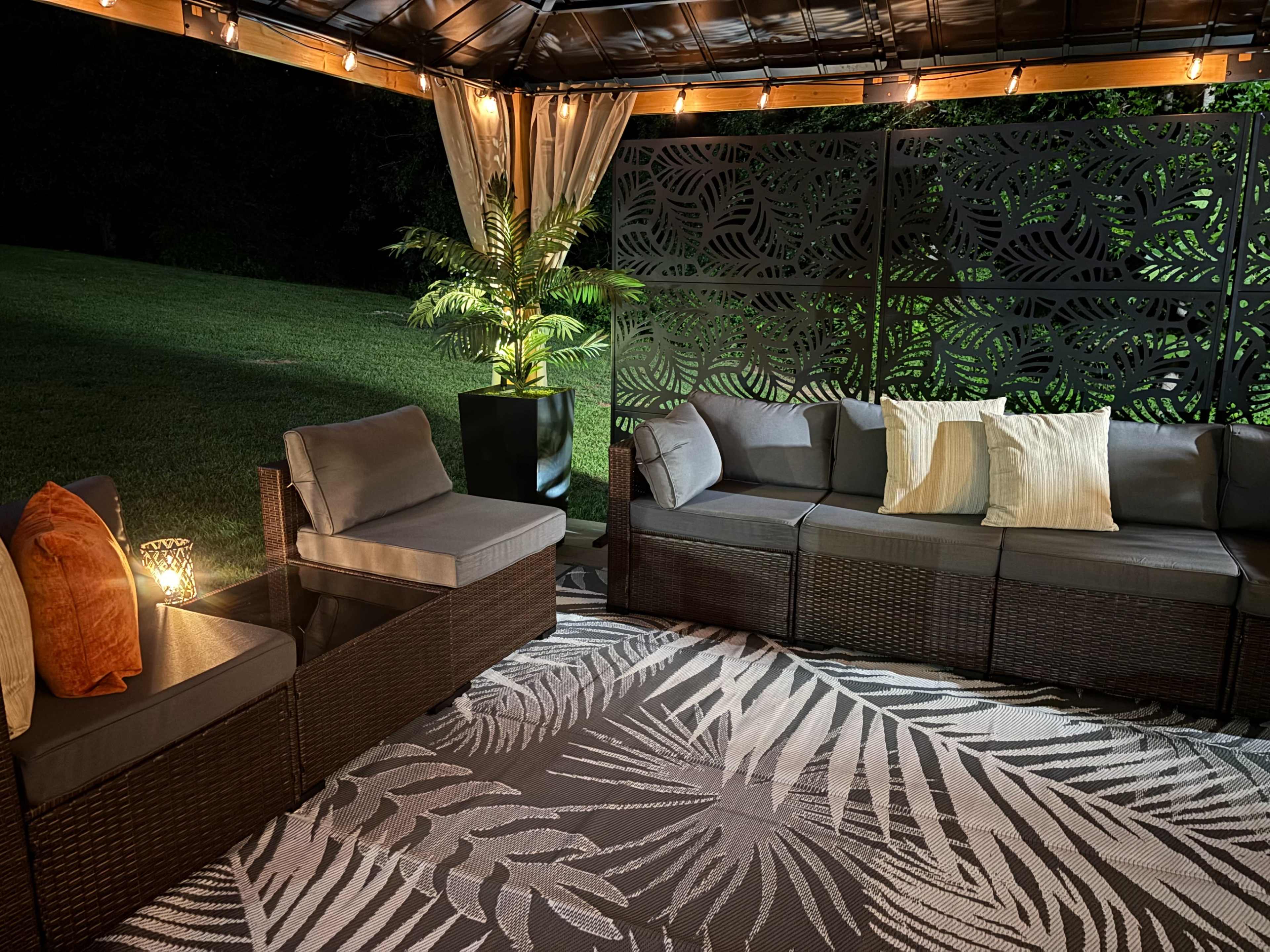 The image shows a patio area with a dark wicker sectional sofa, accent pillows, a glass-topped coffee table, and decorative lights, all under a gazebo surrounded by green grass and a plant.