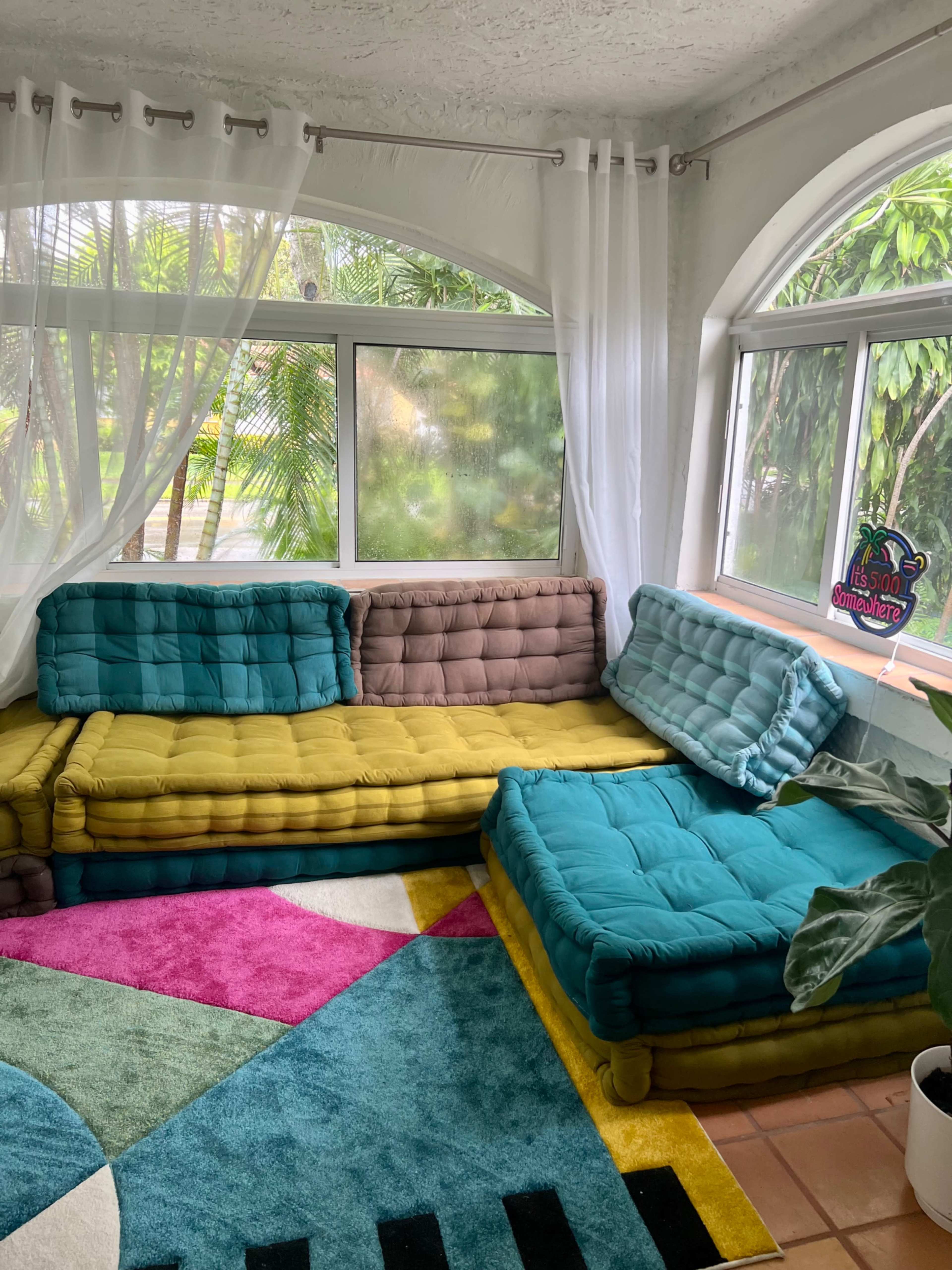 The image shows a colorful seating arrangement with a combination of green, yellow, and blue cushions placed in a sunlit room featuring large windows and greenery outside.