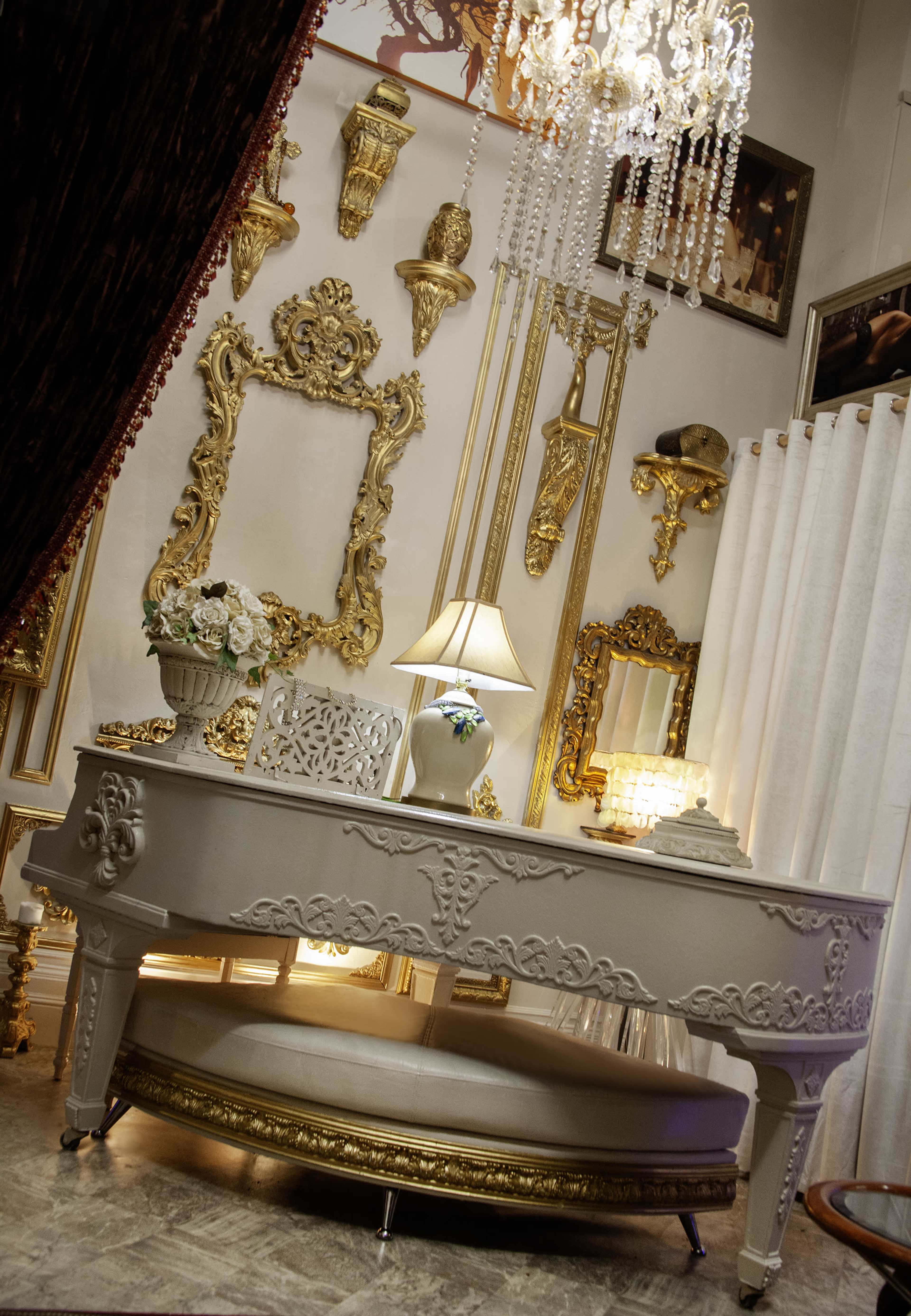The image features an ornate white desk adorned with intricate carvings, set against a backdrop of gold-framed mirrors and decorative wall accents in a lavishly styled room.