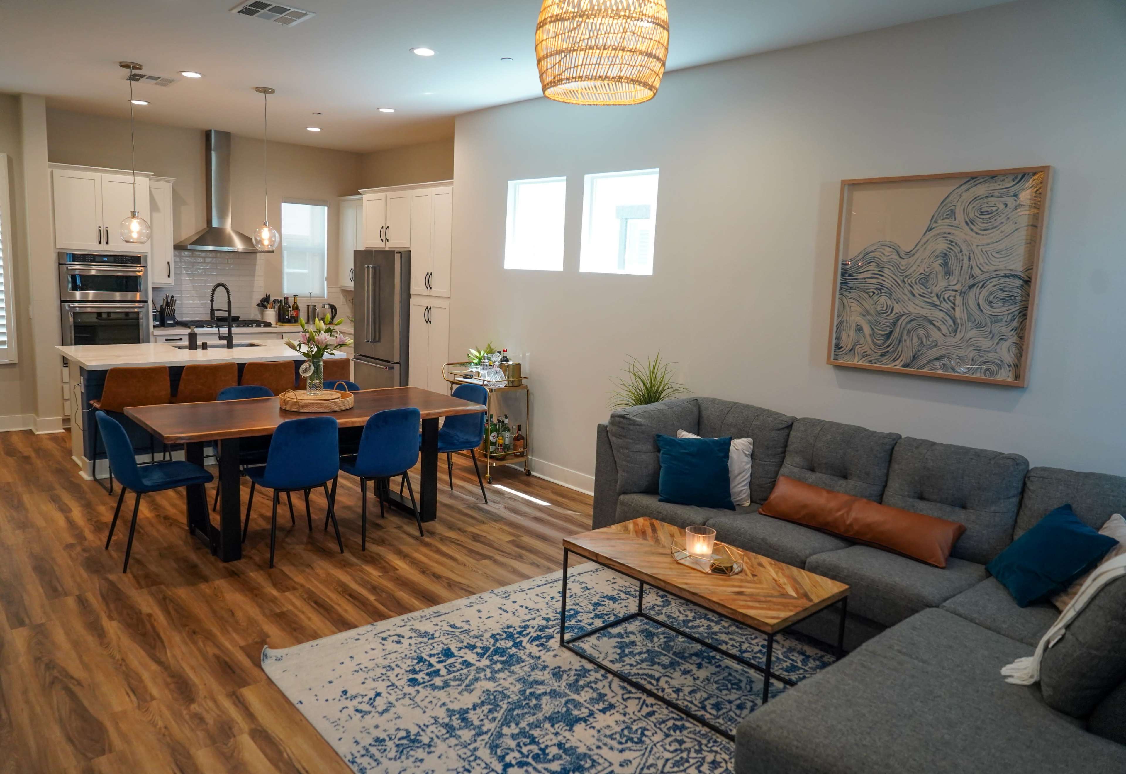 A modern living and dining area features a gray sectional sofa, a wooden dining table with blue chairs, and a kitchen in the background.