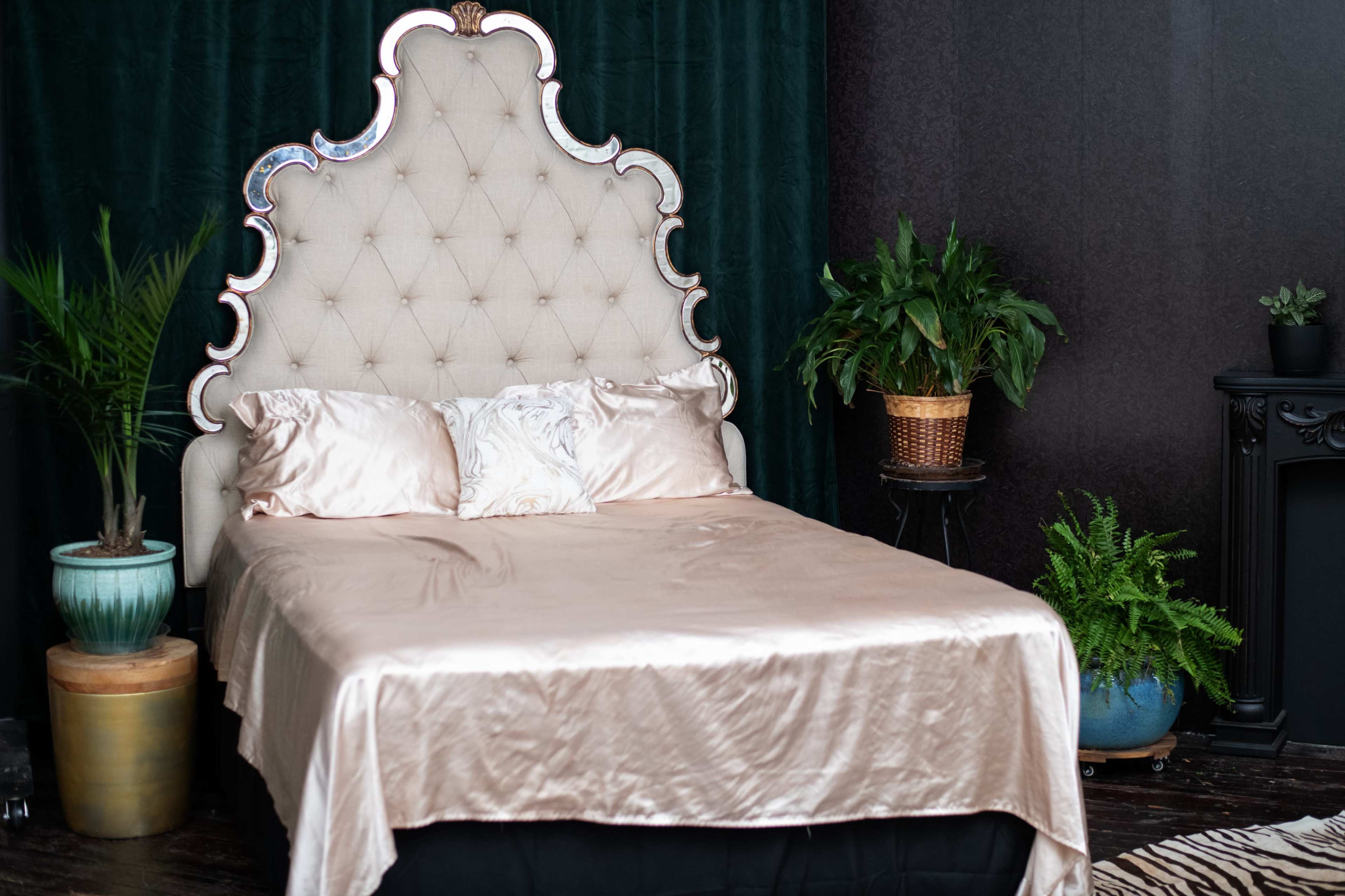 A tufted beige bed with a large, ornate headboard is positioned against a dark wall, surrounded by potted plants.