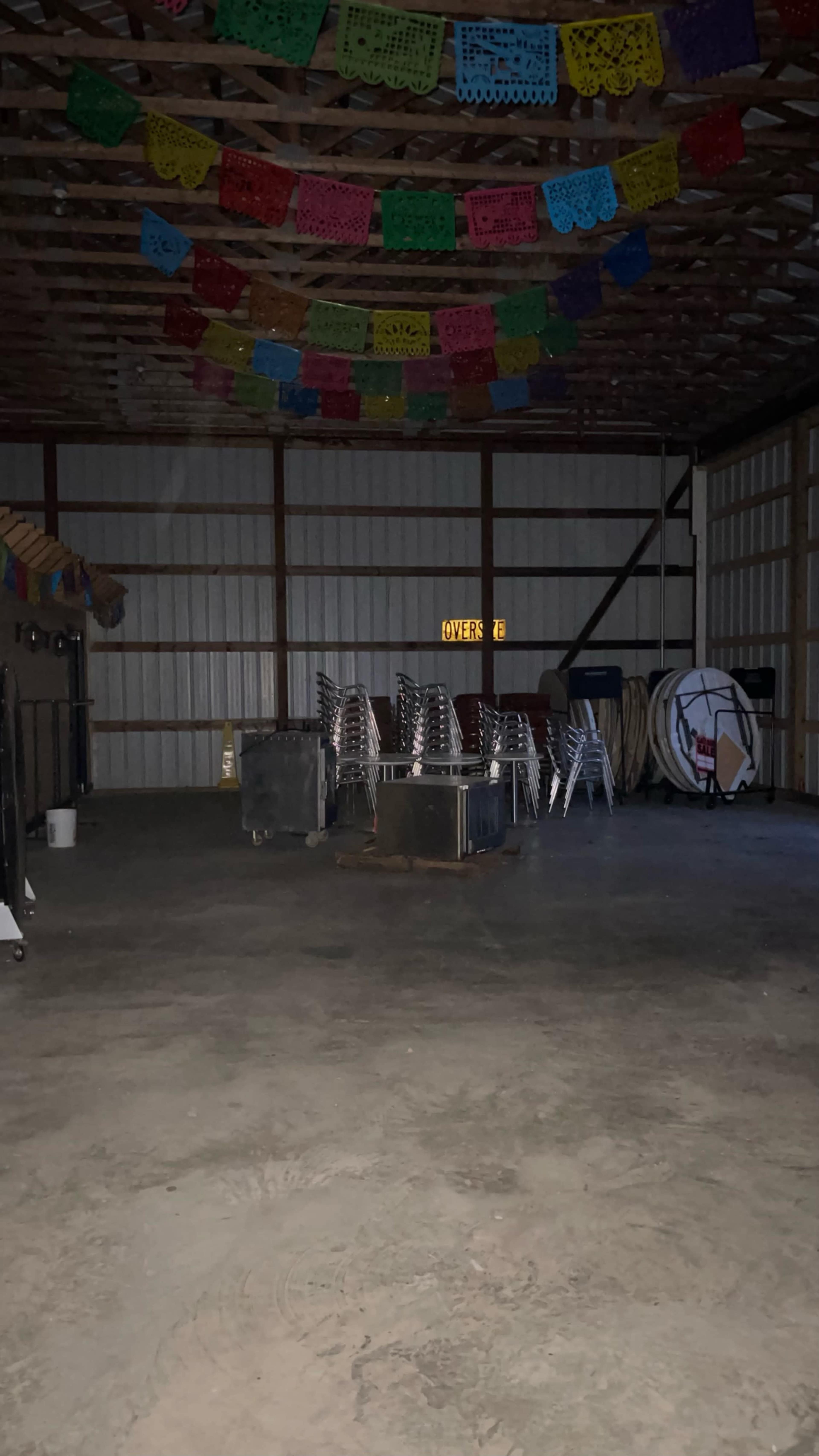 A dimly lit indoor space with metal chairs stacked in the center and colorful papel picado banners hanging from the ceiling.