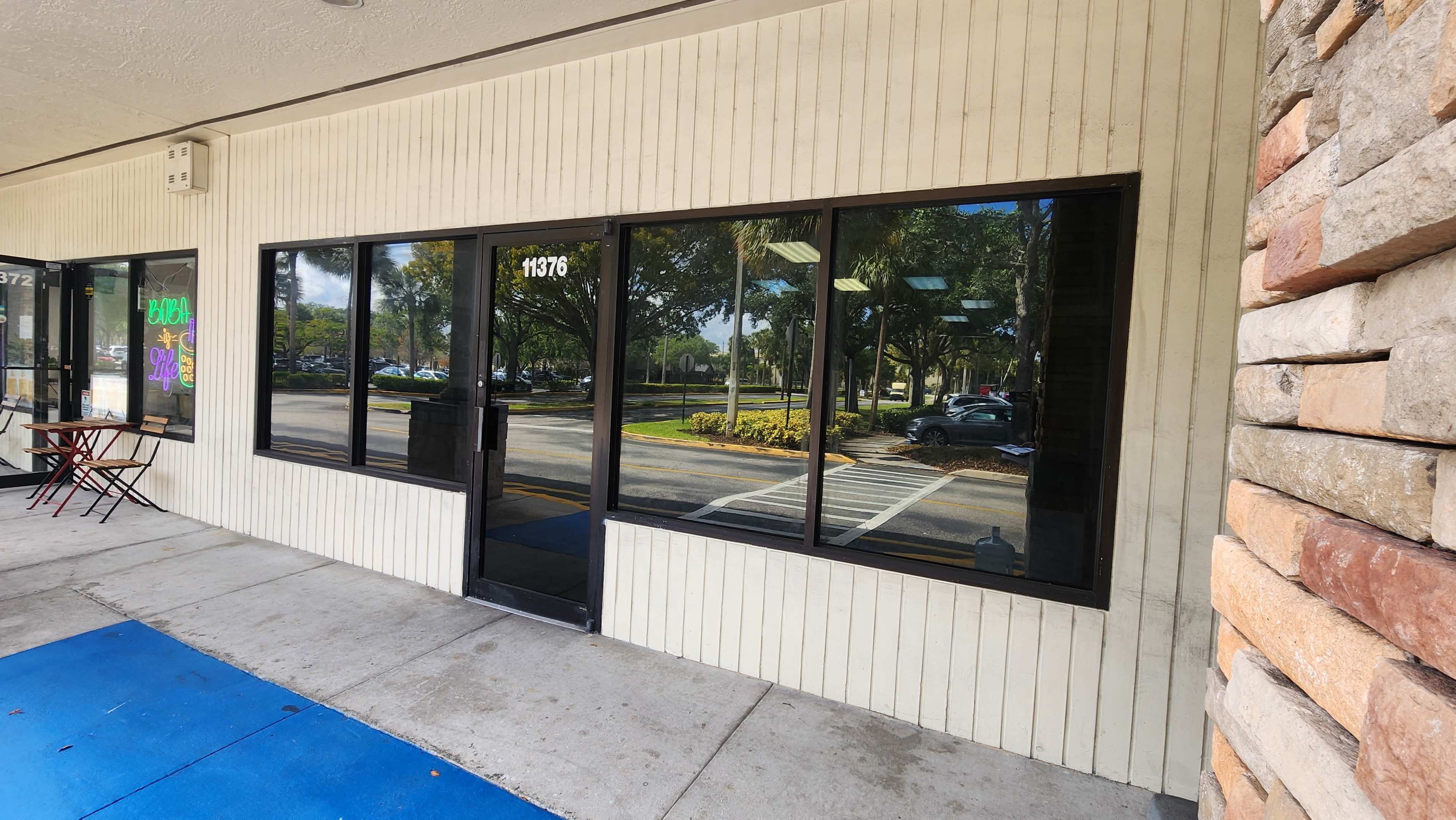 The image shows the exterior of a commercial building featuring large glass windows, a black door, and a section of stone siding next to a sidewalk.