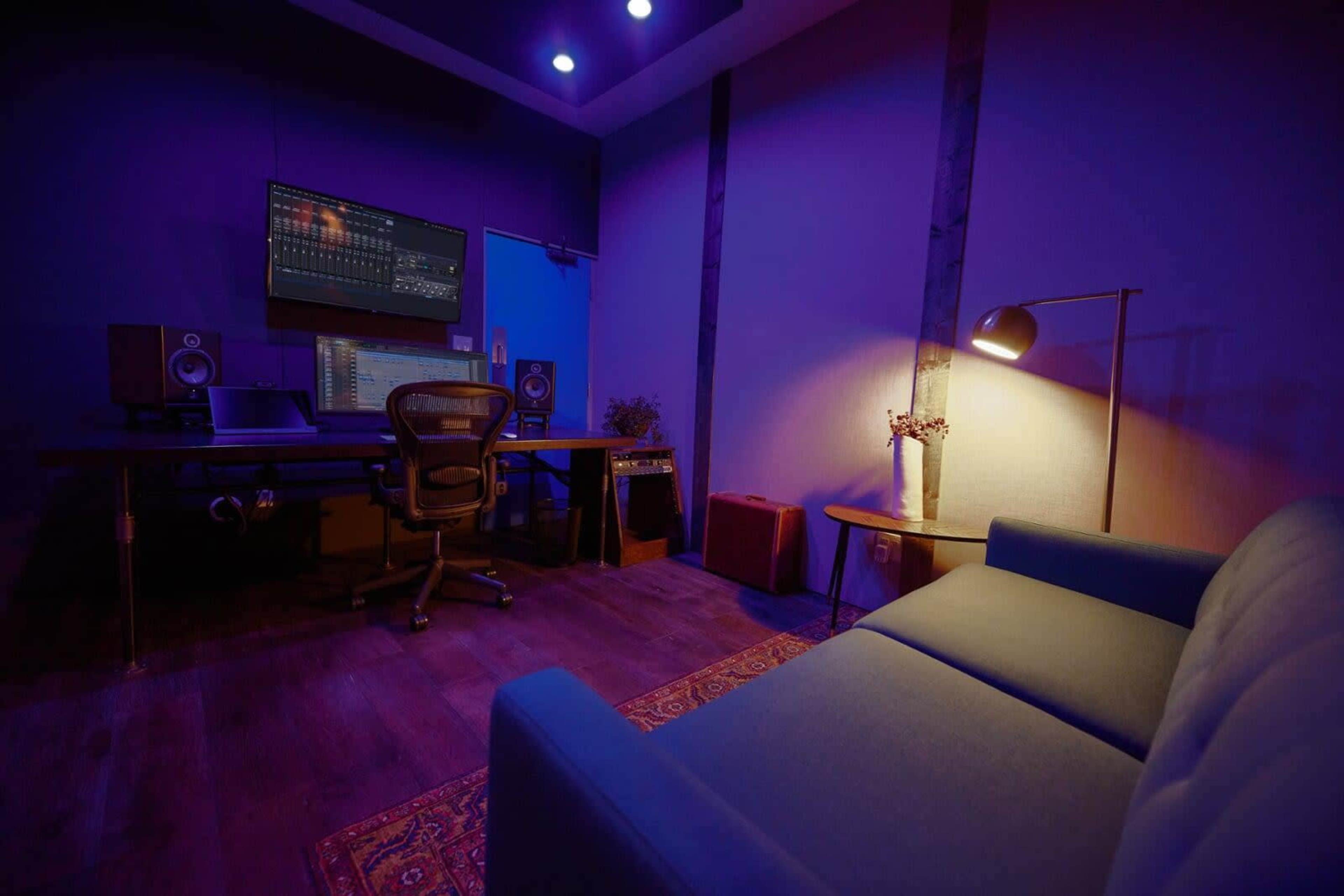 The image shows a modern recording studio with a desk, computer equipment, speakers, a comfortable chair, and a sofa under soft lighting.