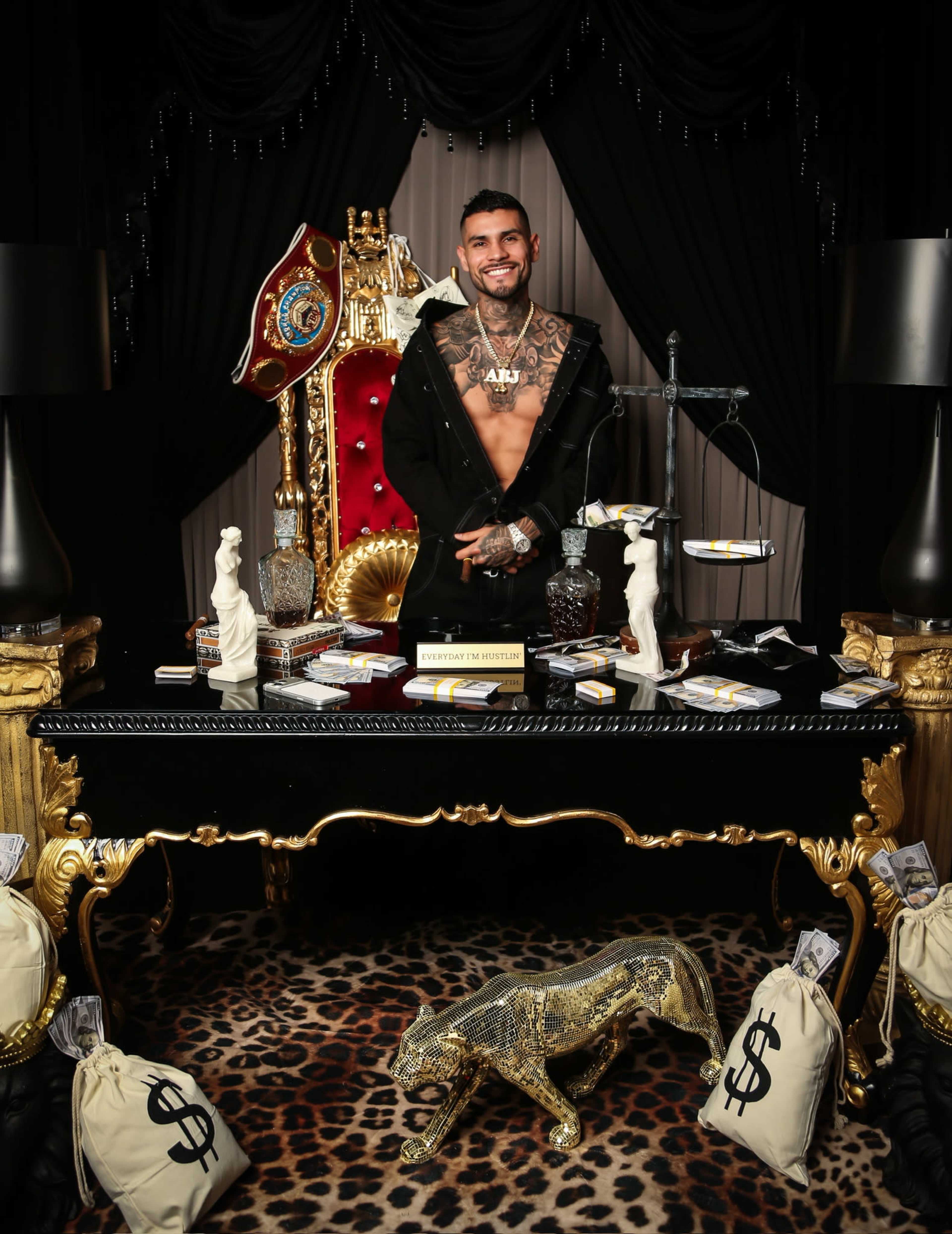 A man with tattoos stands in front of a lavish desk adorned with cash bags, figurines, and an ornate throne in a dark room with elaborate decor.