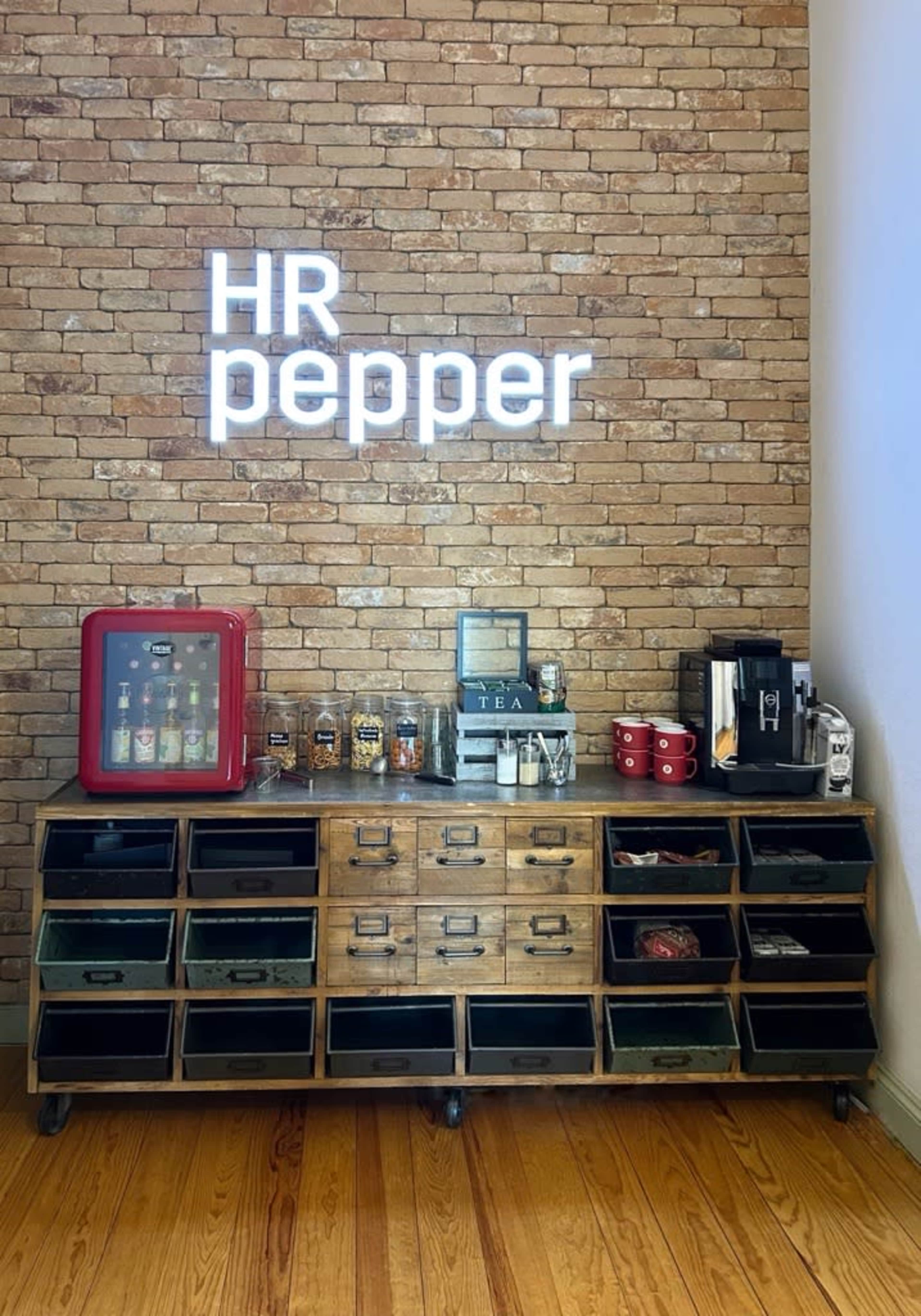 The image shows a wooden storage unit on wheels against a brick wall, with a neon sign reading "HR pepper" and various beverage containers on top.