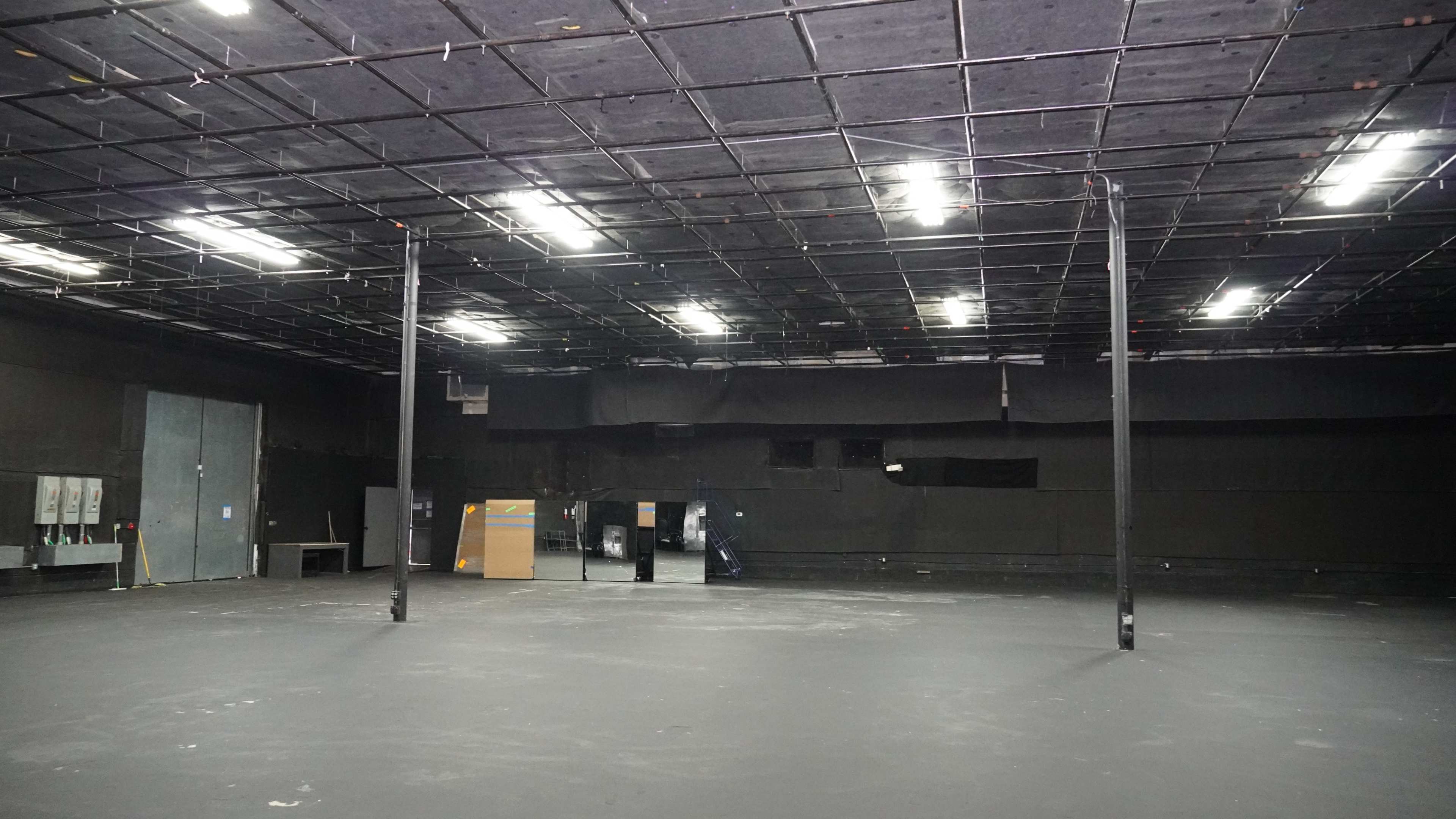 The image depicts a large, empty studio space with a dark ceiling and walls, featuring a smooth black floor and a few visible electrical panels.
