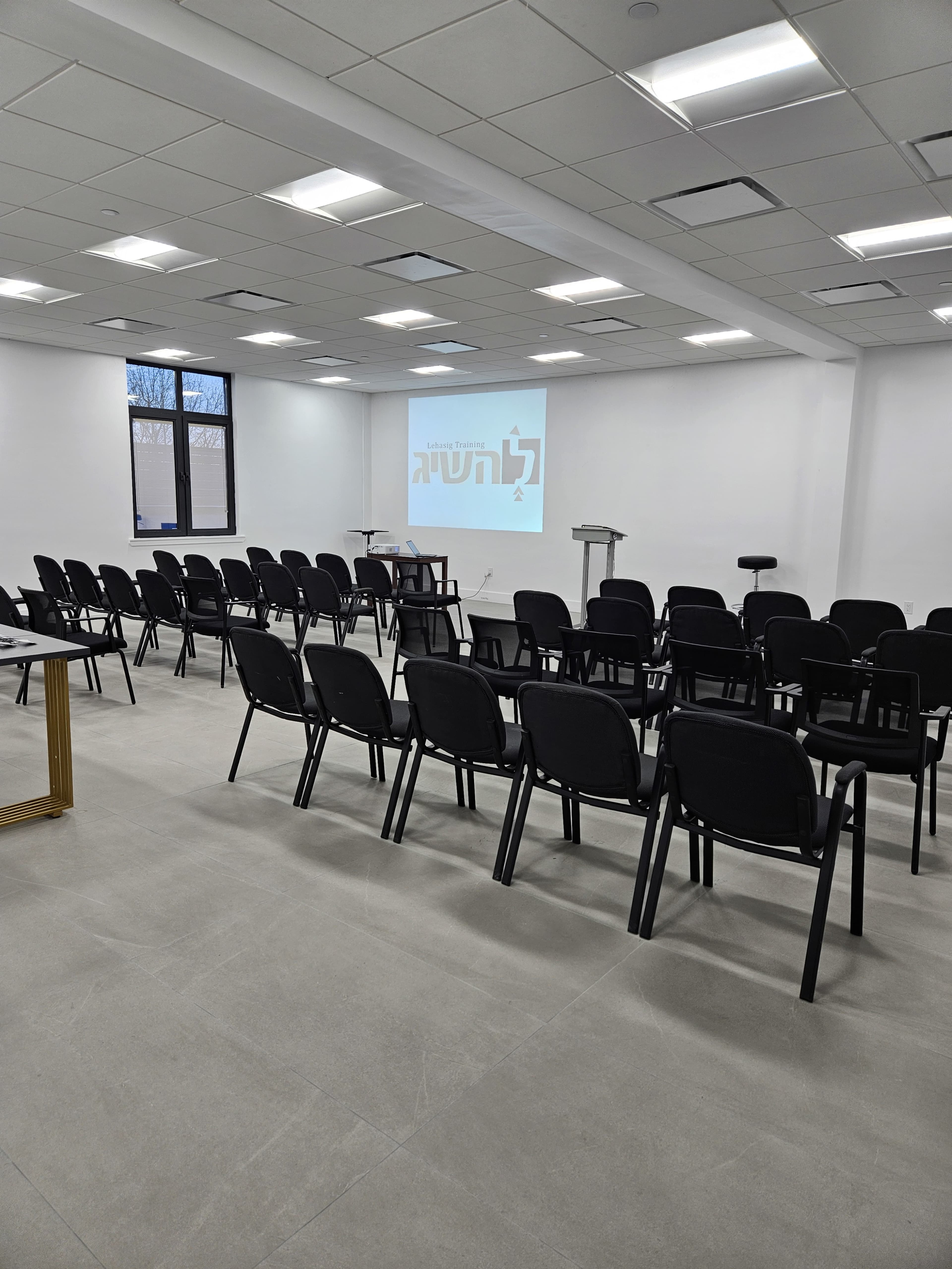 The image shows a conference room with rows of black chairs facing a projector screen displaying Hebrew text.