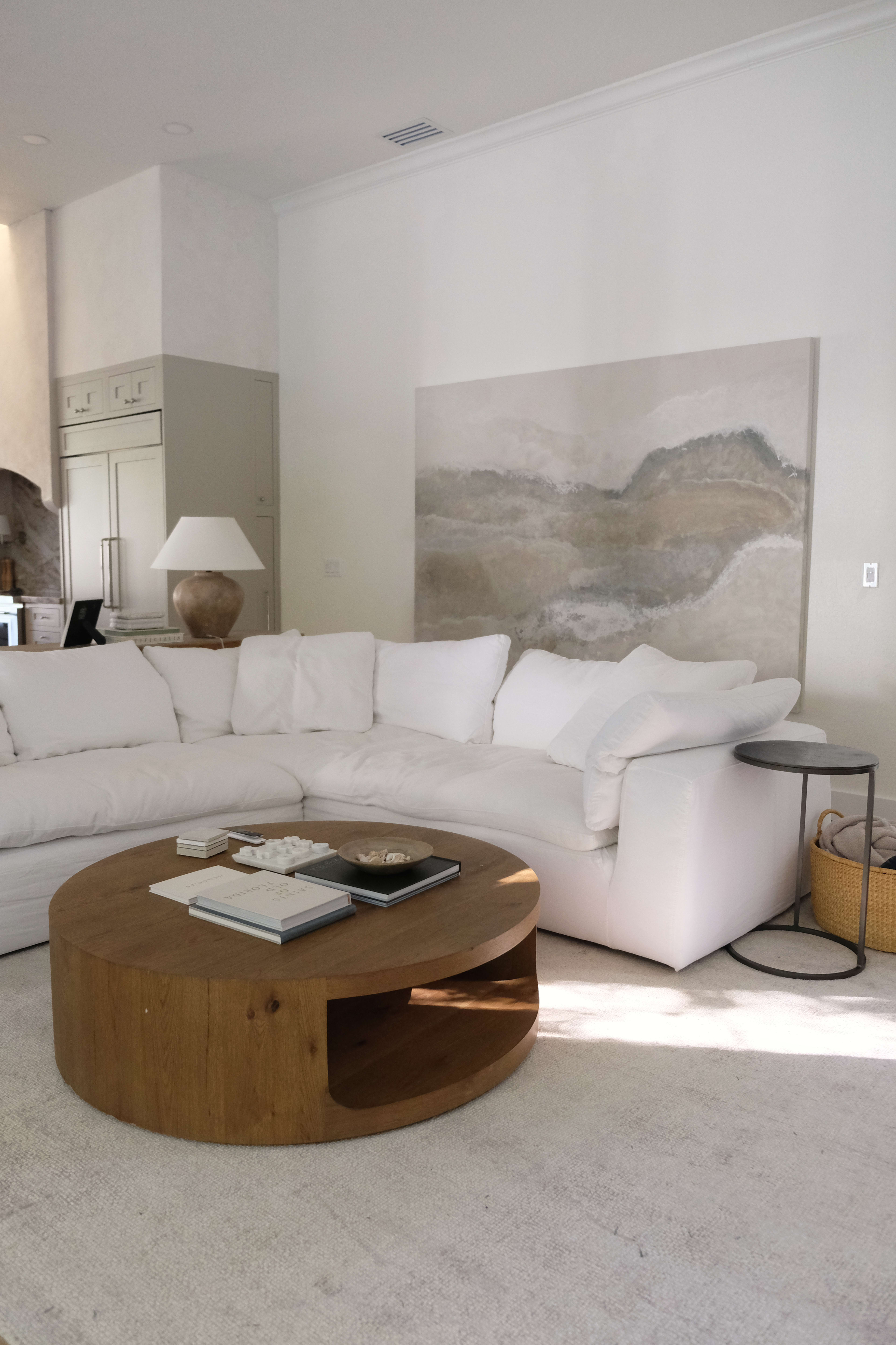 The image shows a modern living room featuring a curved white sectional sofa, a round wooden coffee table, and a large abstract painting on the wall.