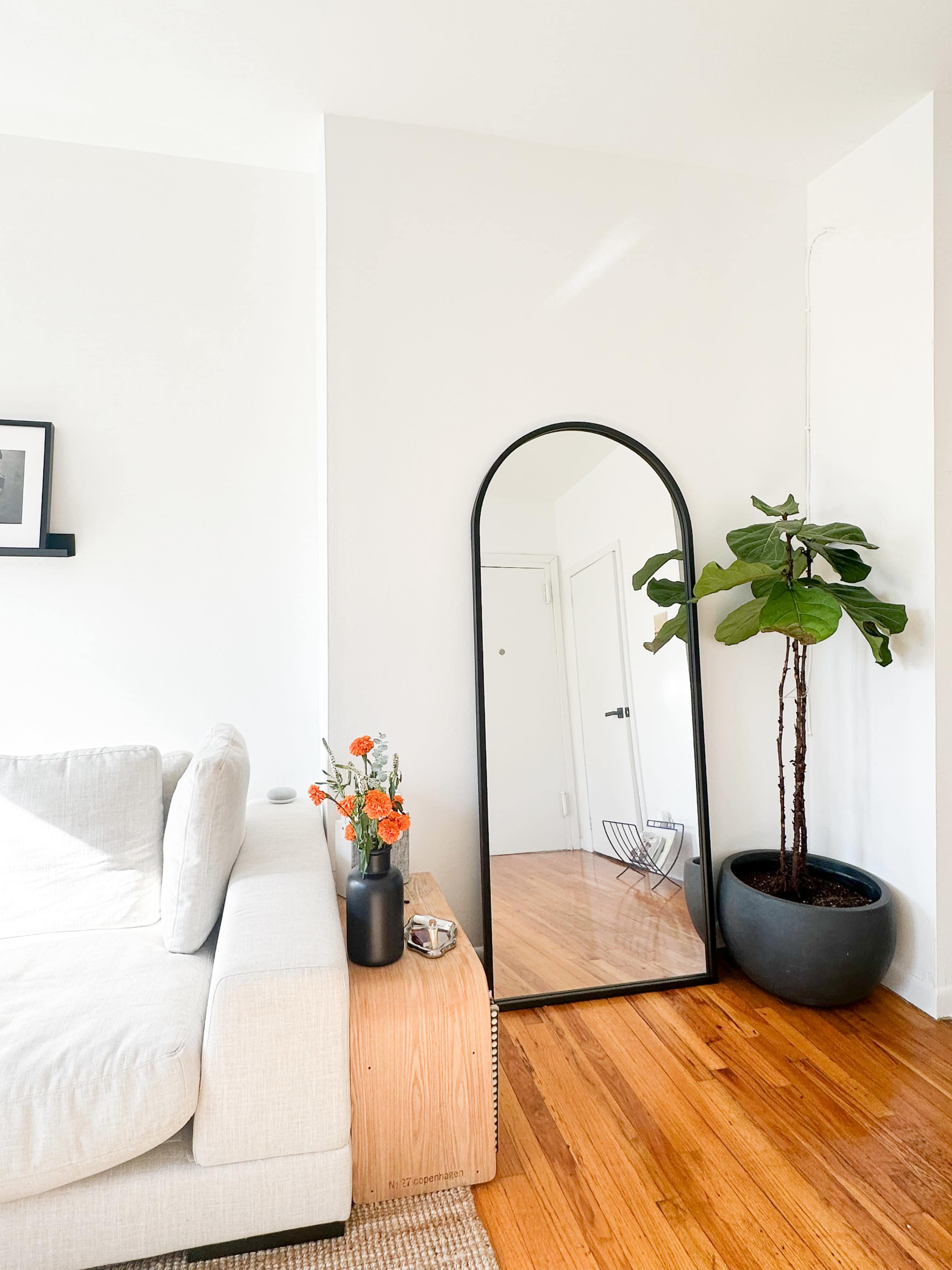 A tall, arched mirror stands beside a potted plant in a bright corner of a living room, next to a light-colored sofa and a small vase of flowers.