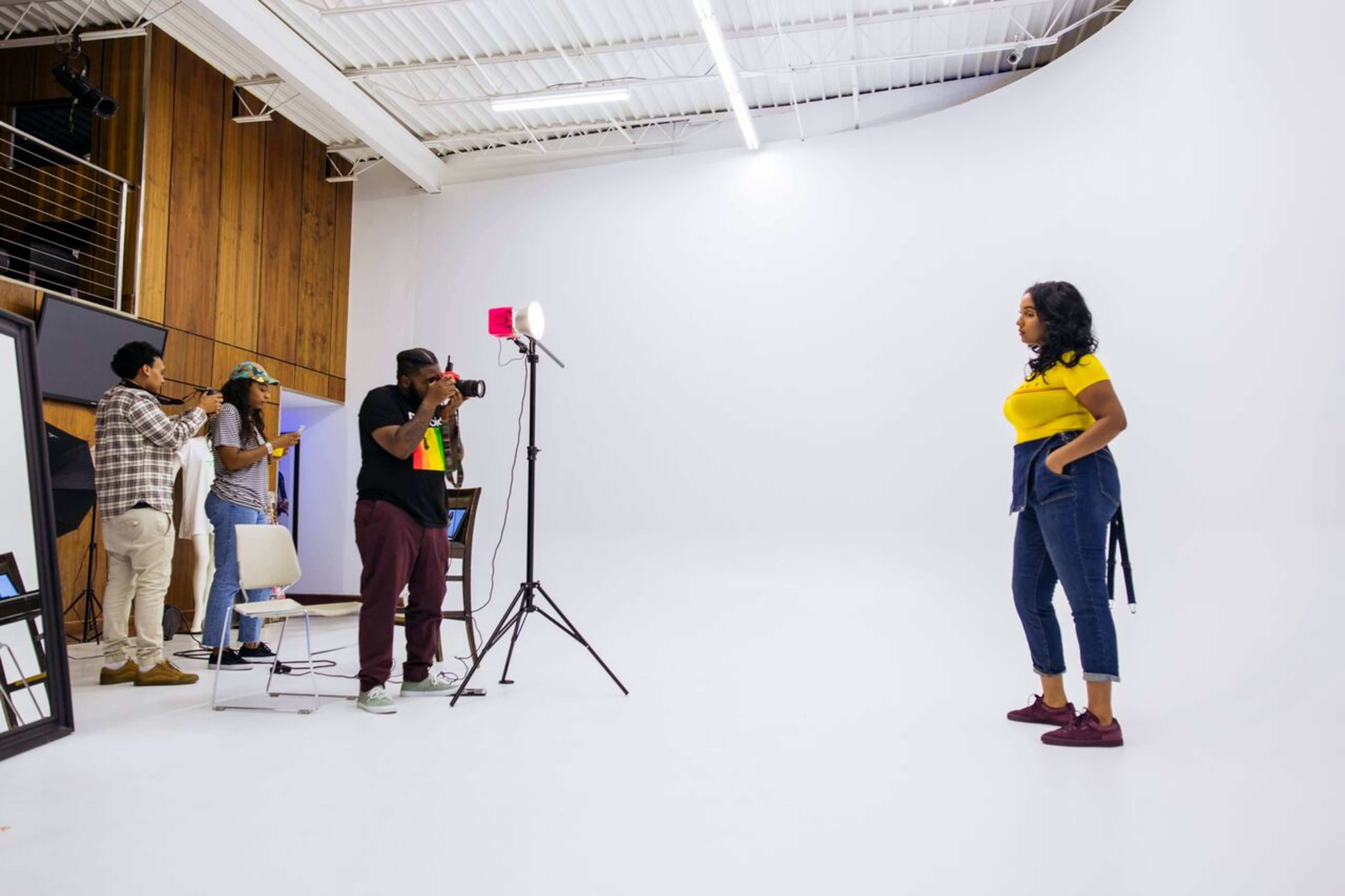 A photoshoot is taking place in a studio with a model posing against a white backdrop while photographers and crew are set up to capture the images.
