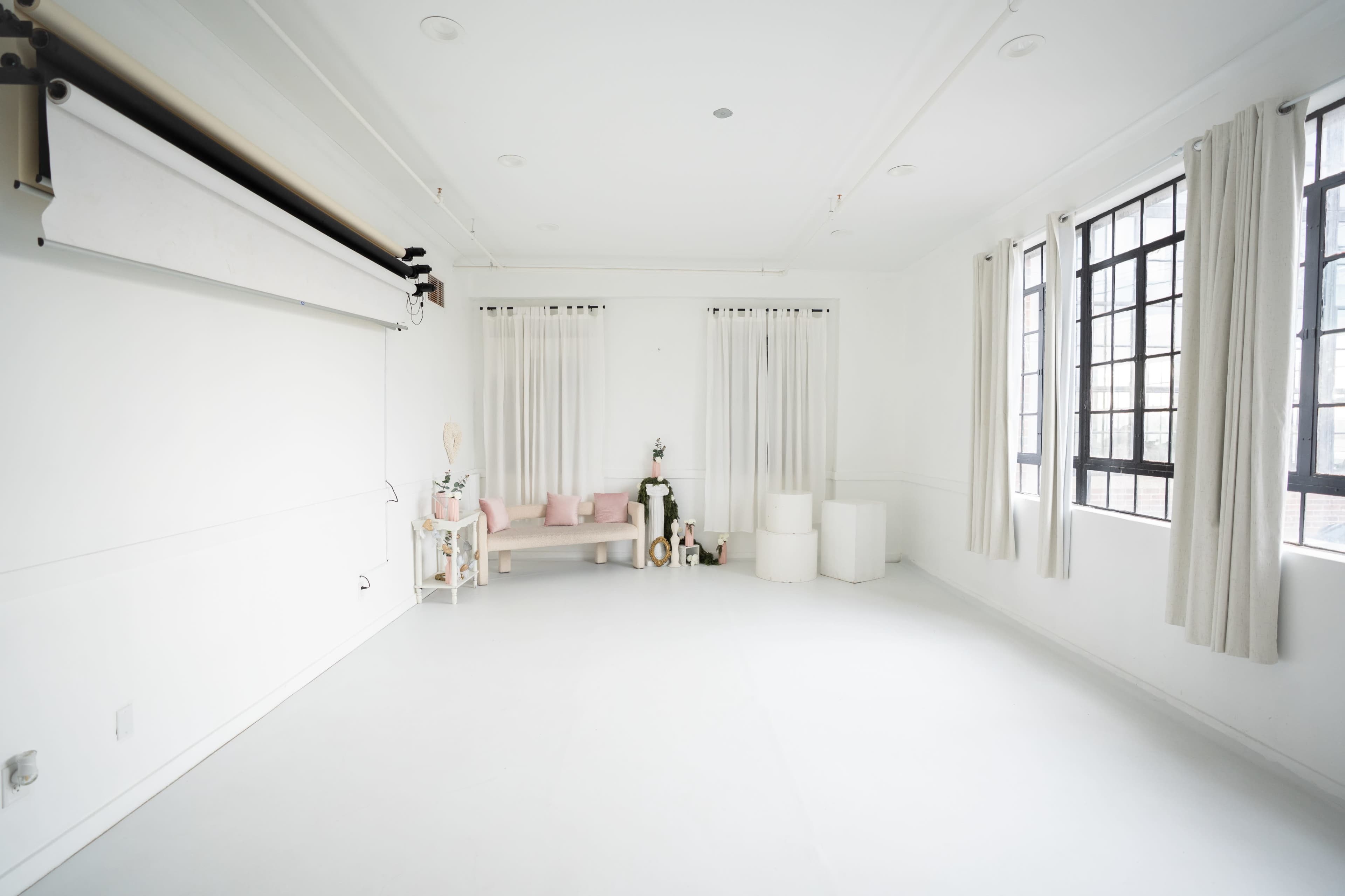The image shows a bright, minimalist studio space with white walls, large windows, and a simple seating arrangement.