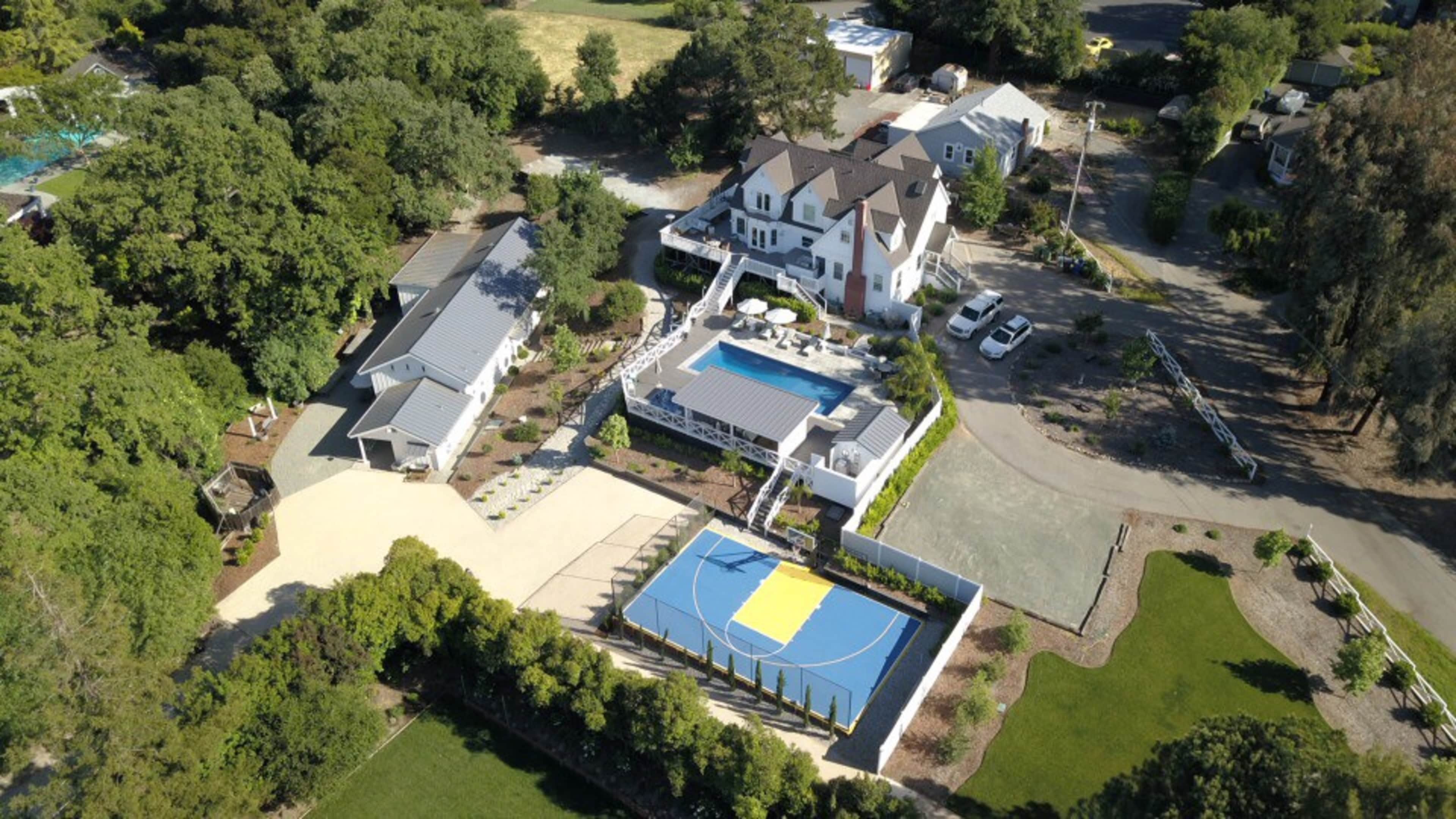 The aerial view shows a large residential property featuring a swimming pool, a basketball court, and multiple structures surrounded by greenery.