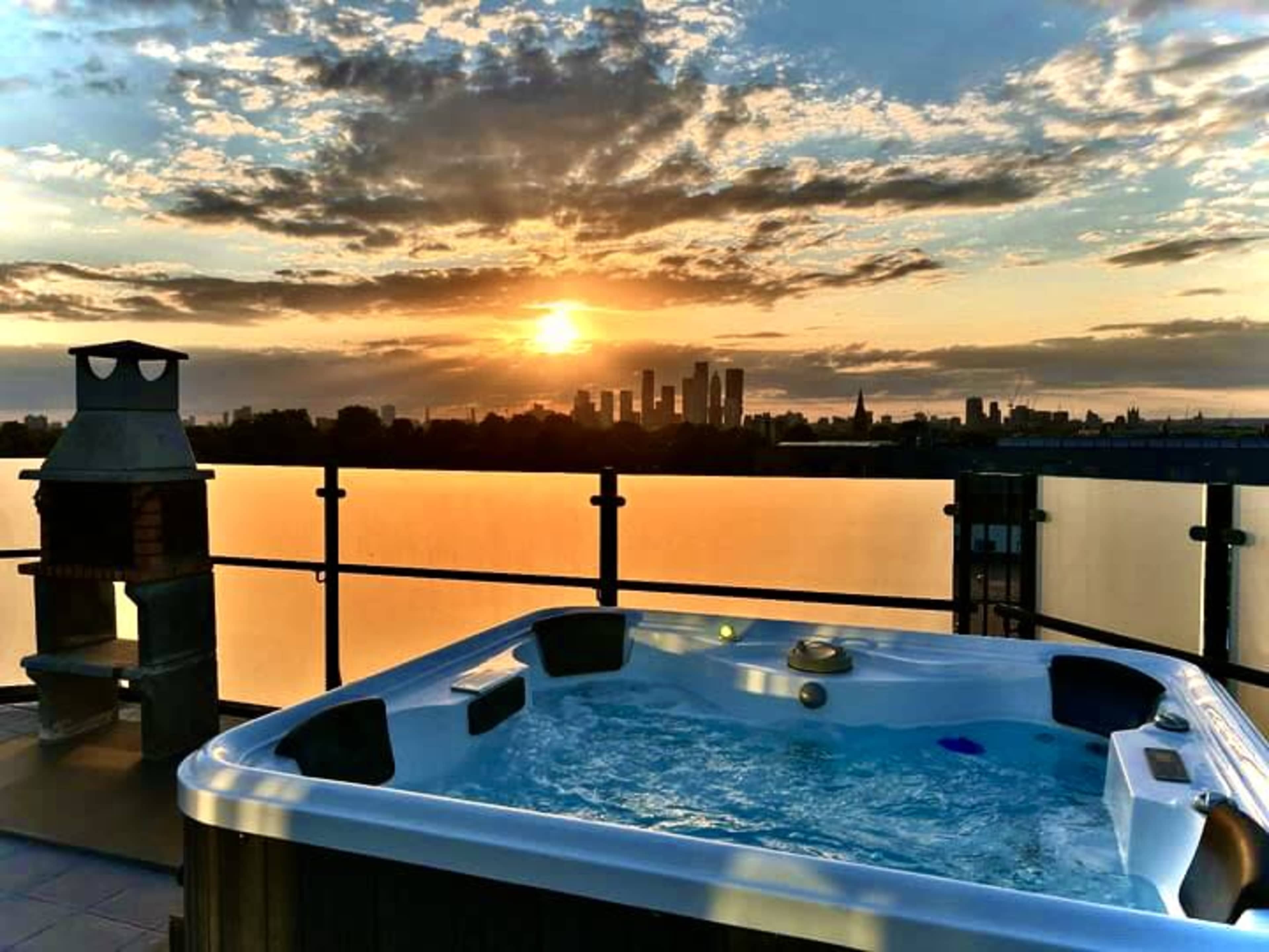 A hot tub is positioned on a rooftop terrace overlooking a city skyline at sunset.