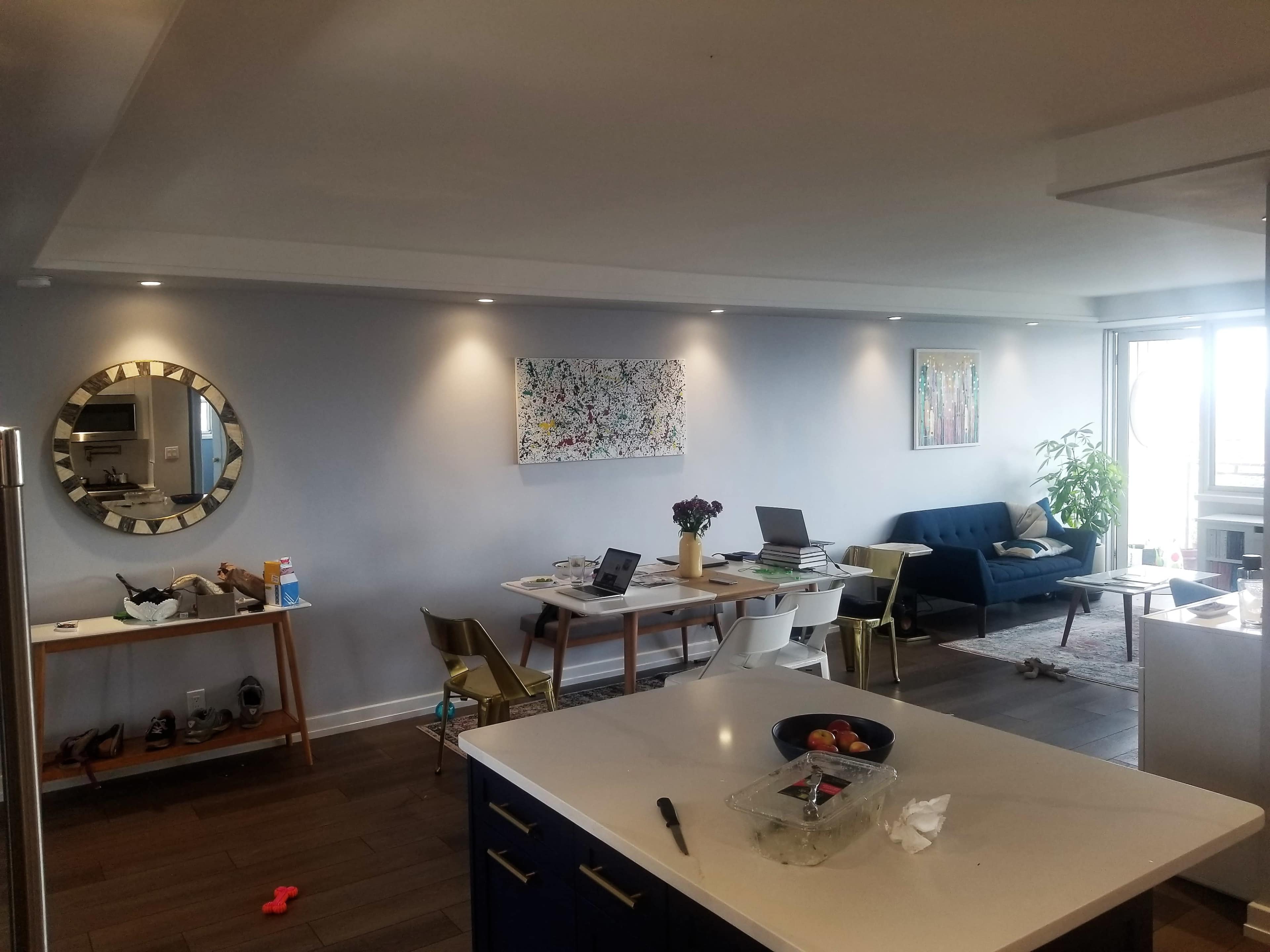 A modern living area features a dining table, a blue couch, and a large window with natural light, alongside decorative artwork and plants.