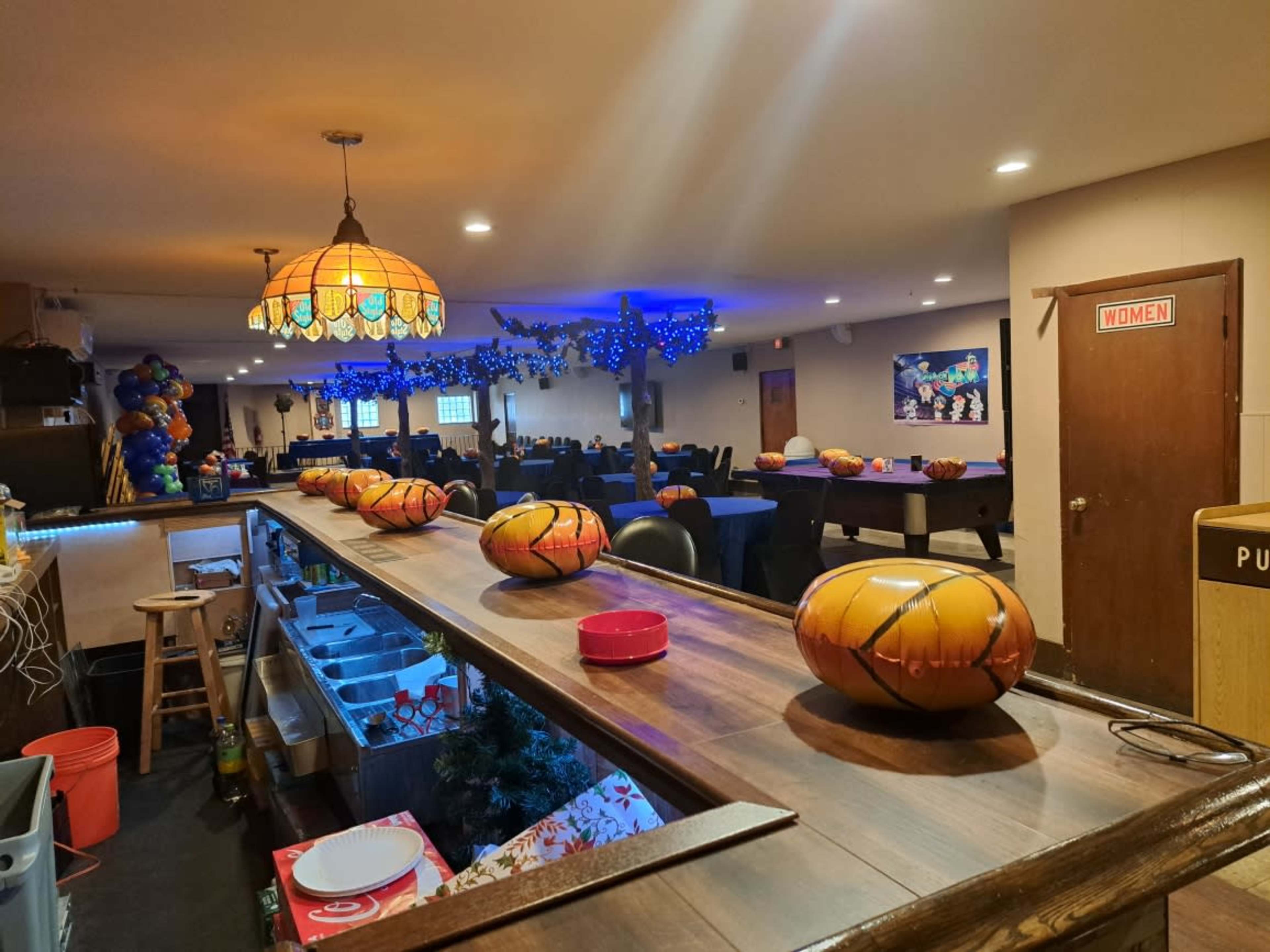 A bar with a polished wooden counter decorated with orange and black pumpkin-shaped items, while the background features tables set up for an event, illuminated by colorful lights.