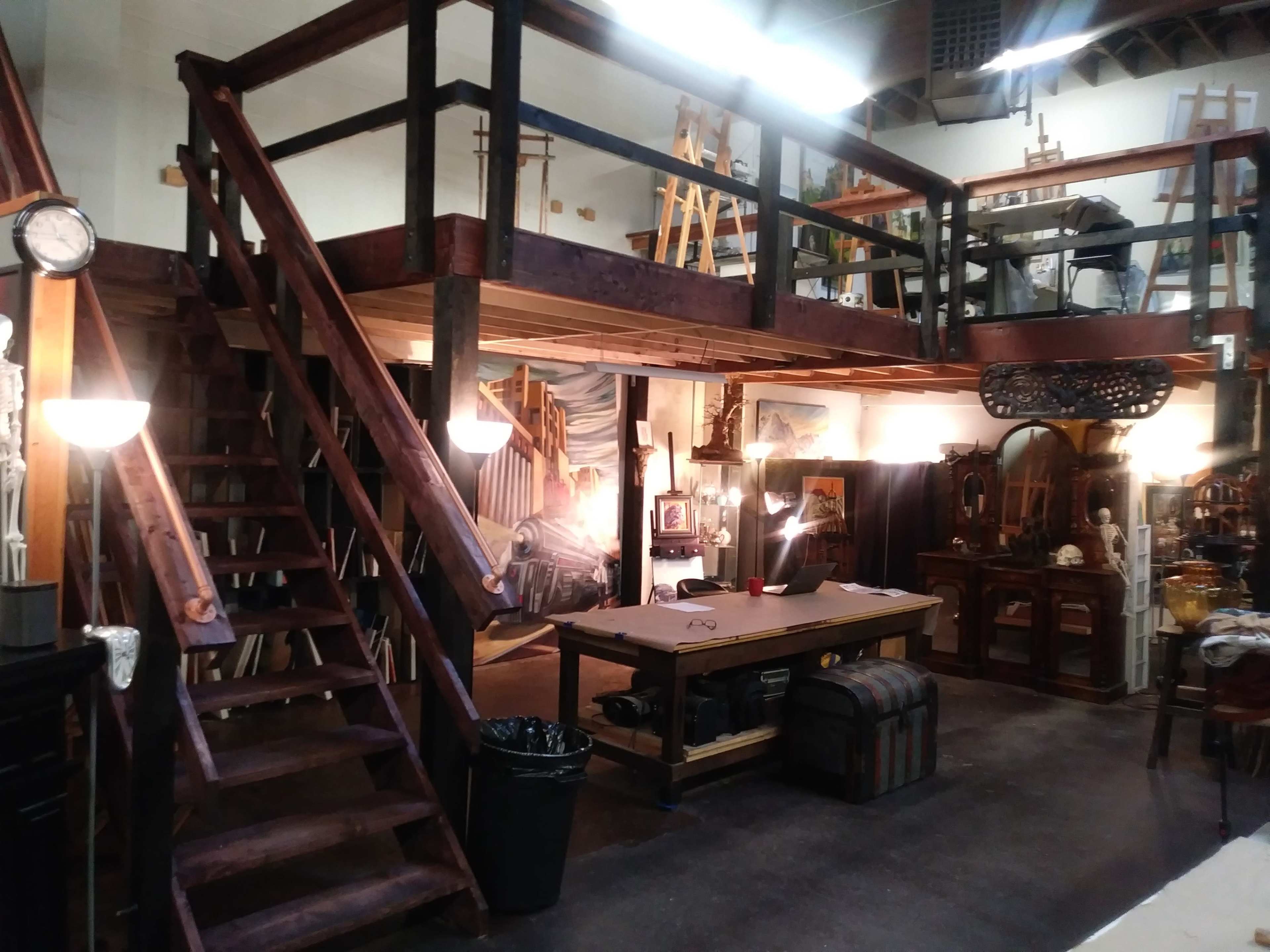 An art studio with a lofted area accessible by a wooden staircase, a central work table, and various art supplies and furniture.