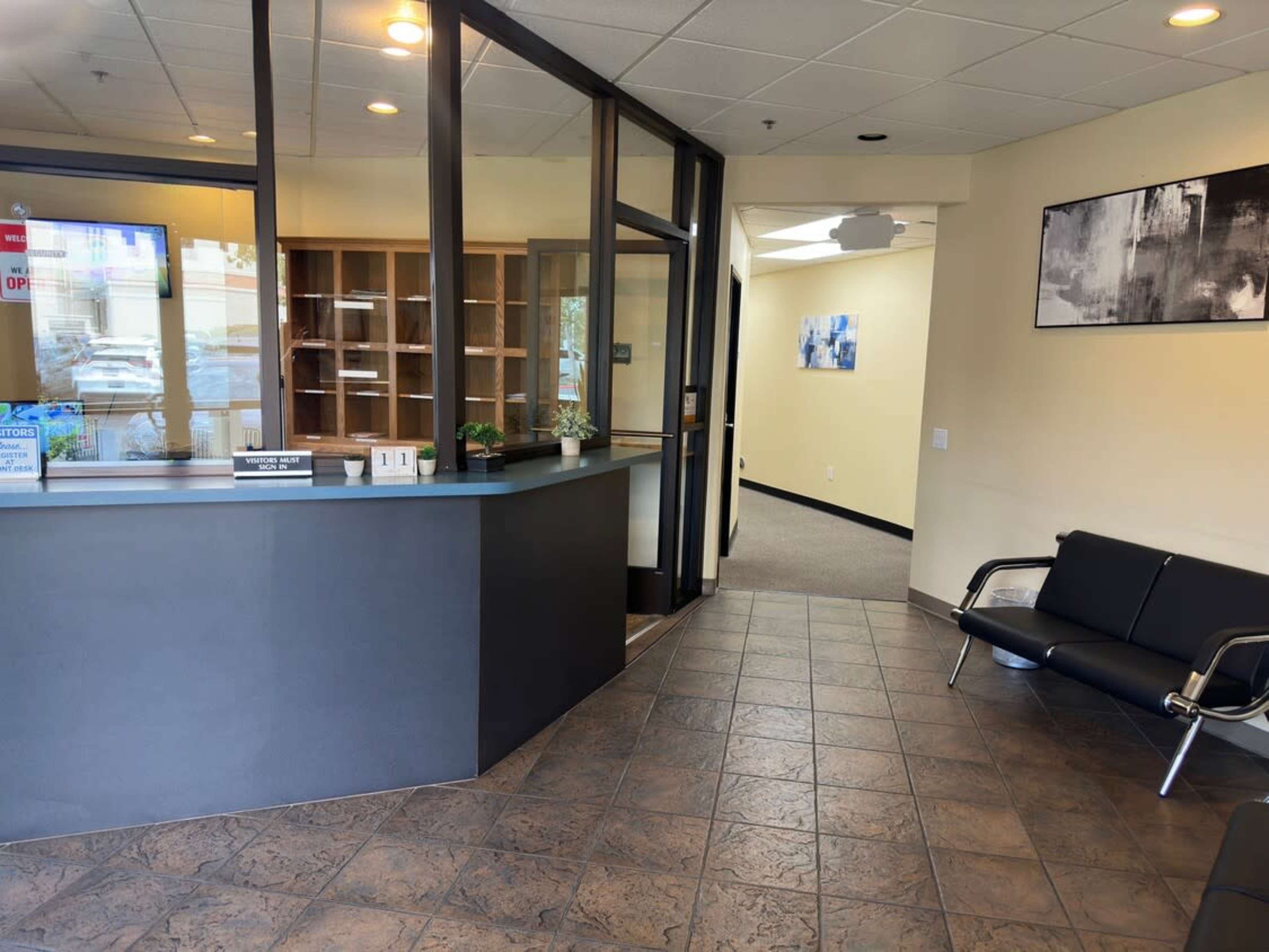 The image shows a modern reception area with a front desk, waiting area seating, and a view into an adjacent room.