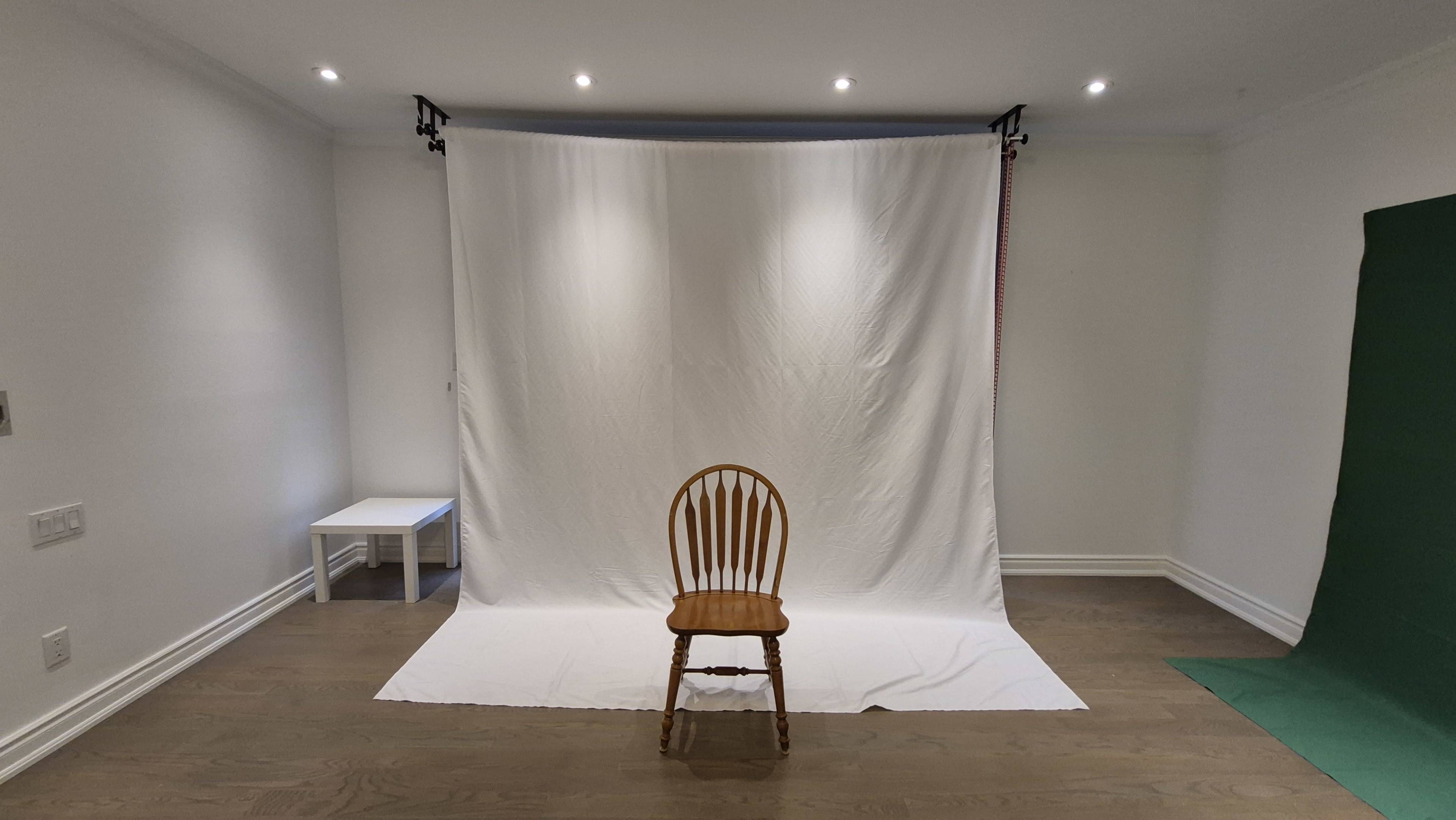 A wooden chair is placed in front of a white backdrop with a green screen to the right in a sparsely furnished room.