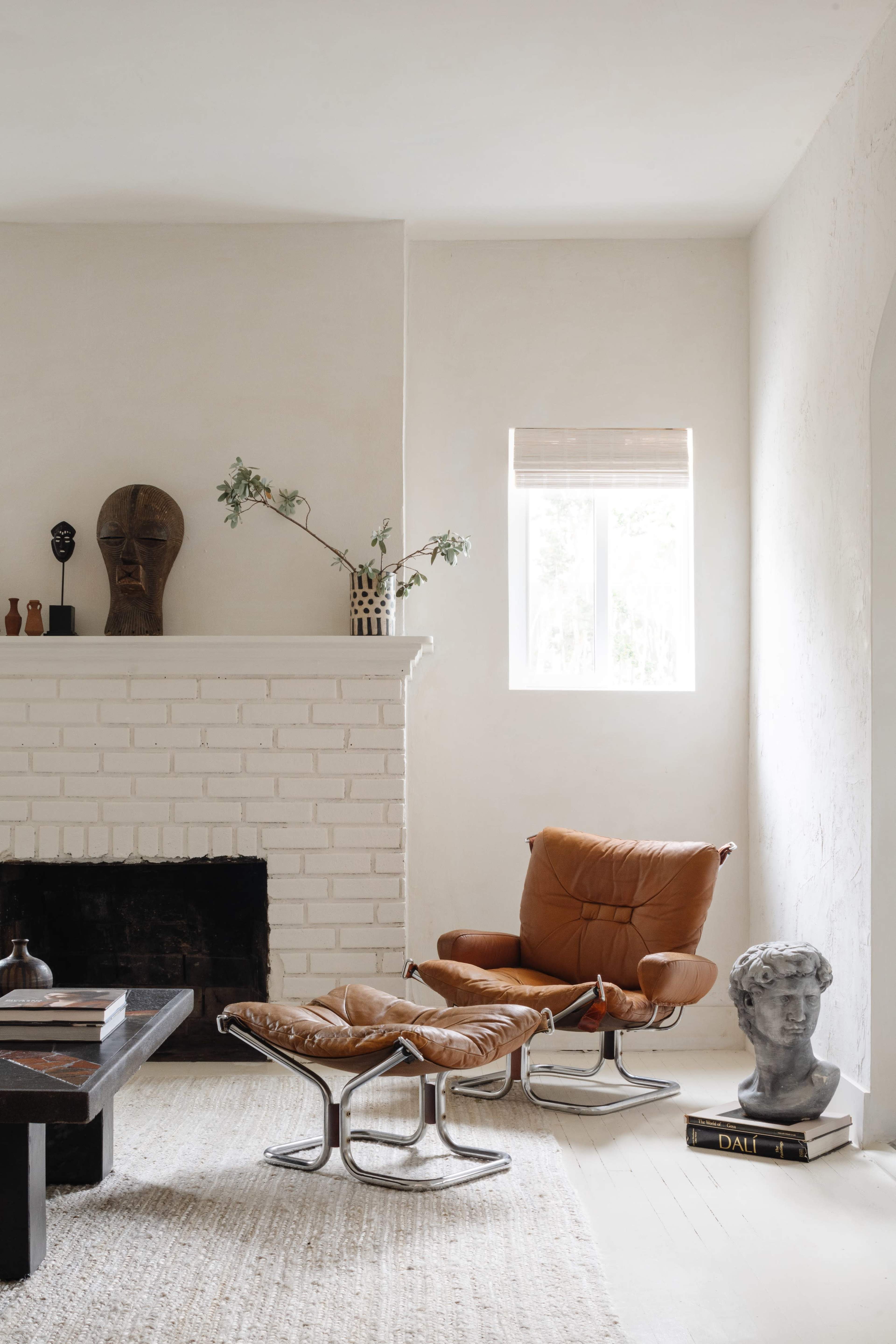 The image features a minimalist living room with a brown leather armchair and ottoman positioned near a white brick fireplace, complemented by a sculpture and decorative items on a shelf.