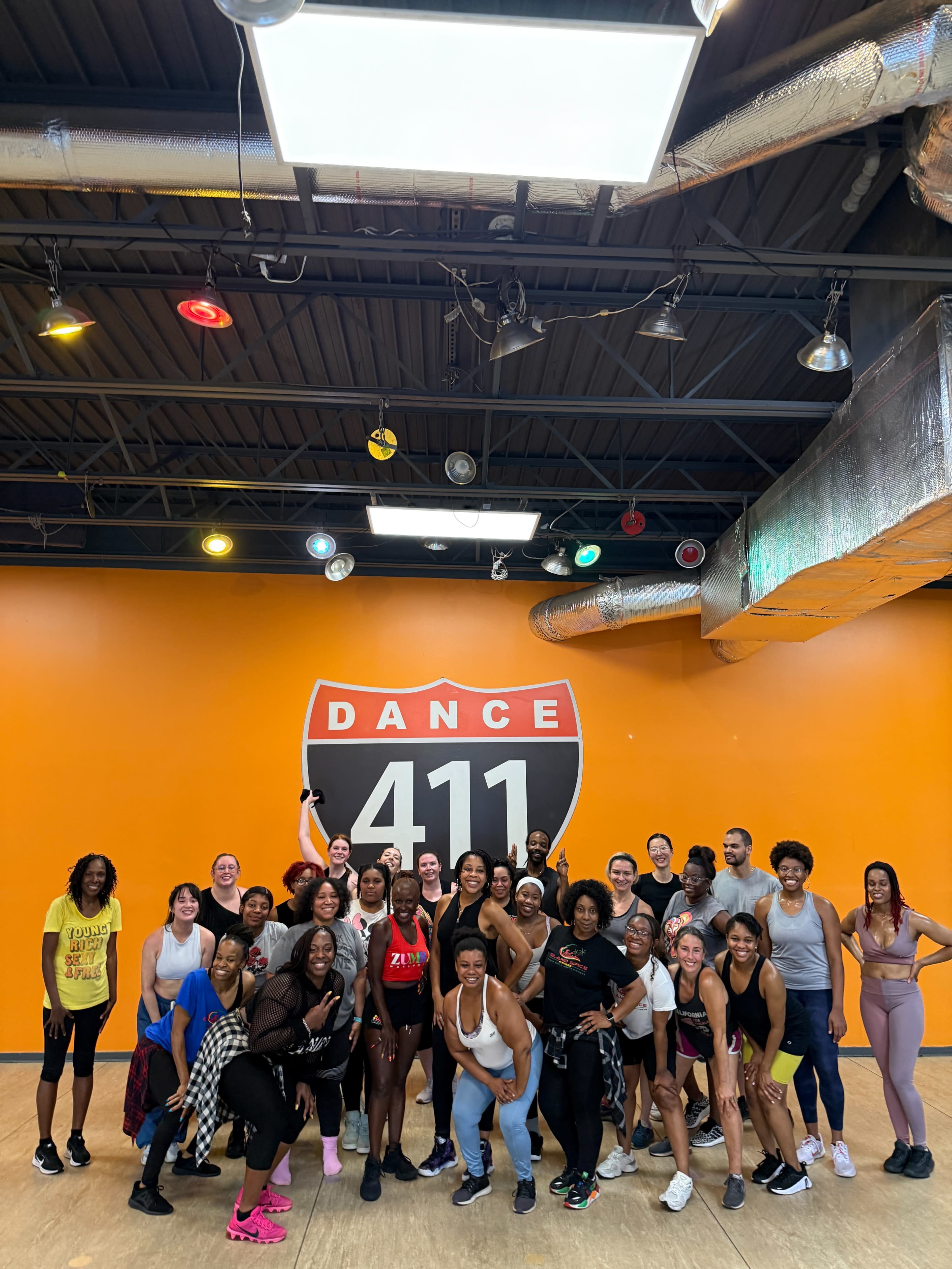 A diverse group of individuals poses together in a dance studio with an orange wall and a sign that says "DANCE 411."