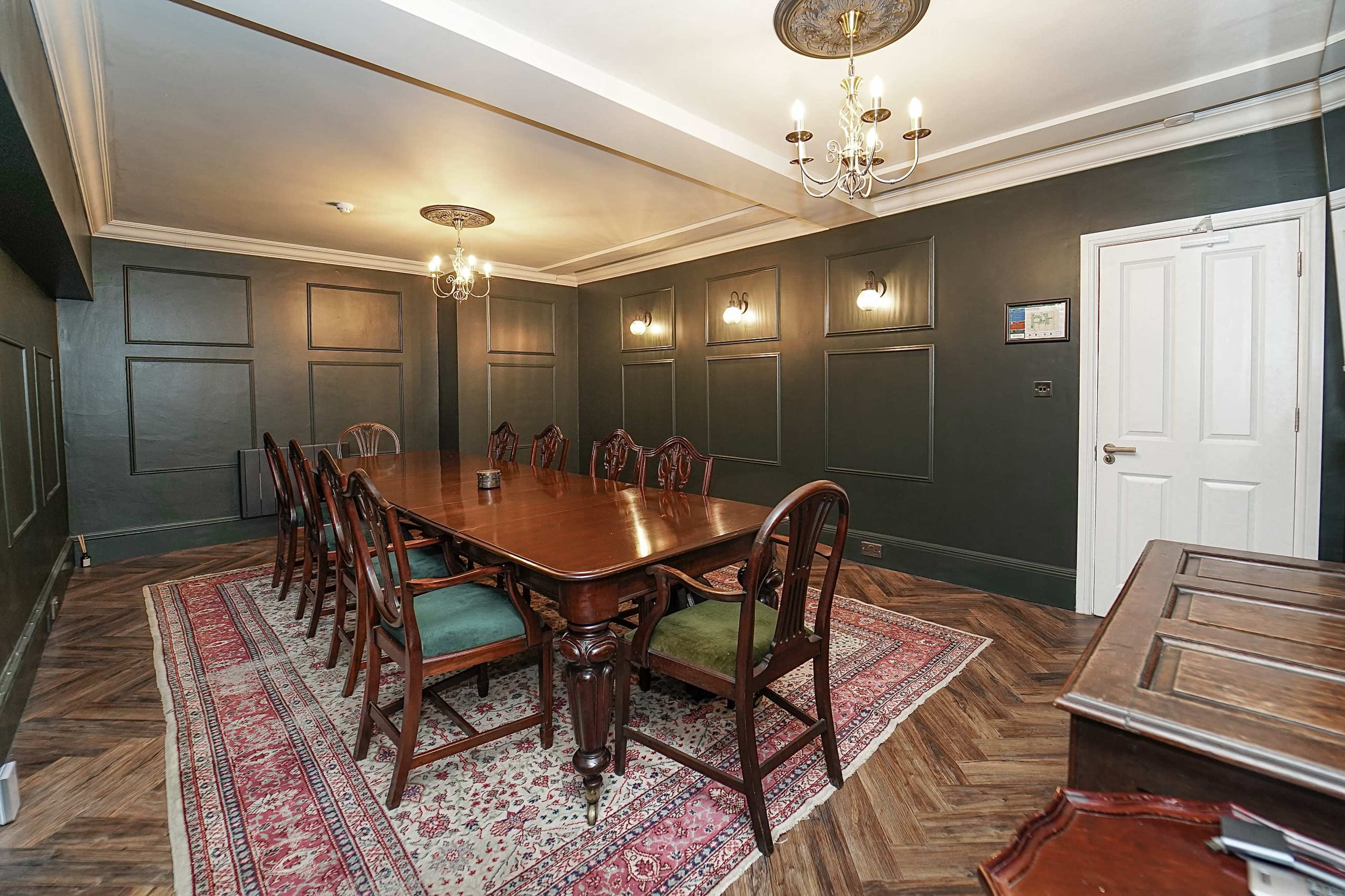 The image shows a formal dining room with a long wooden table surrounded by chairs, a patterned rug on the floor, and decorative wall panels, all under soft lighting from chandeliers.