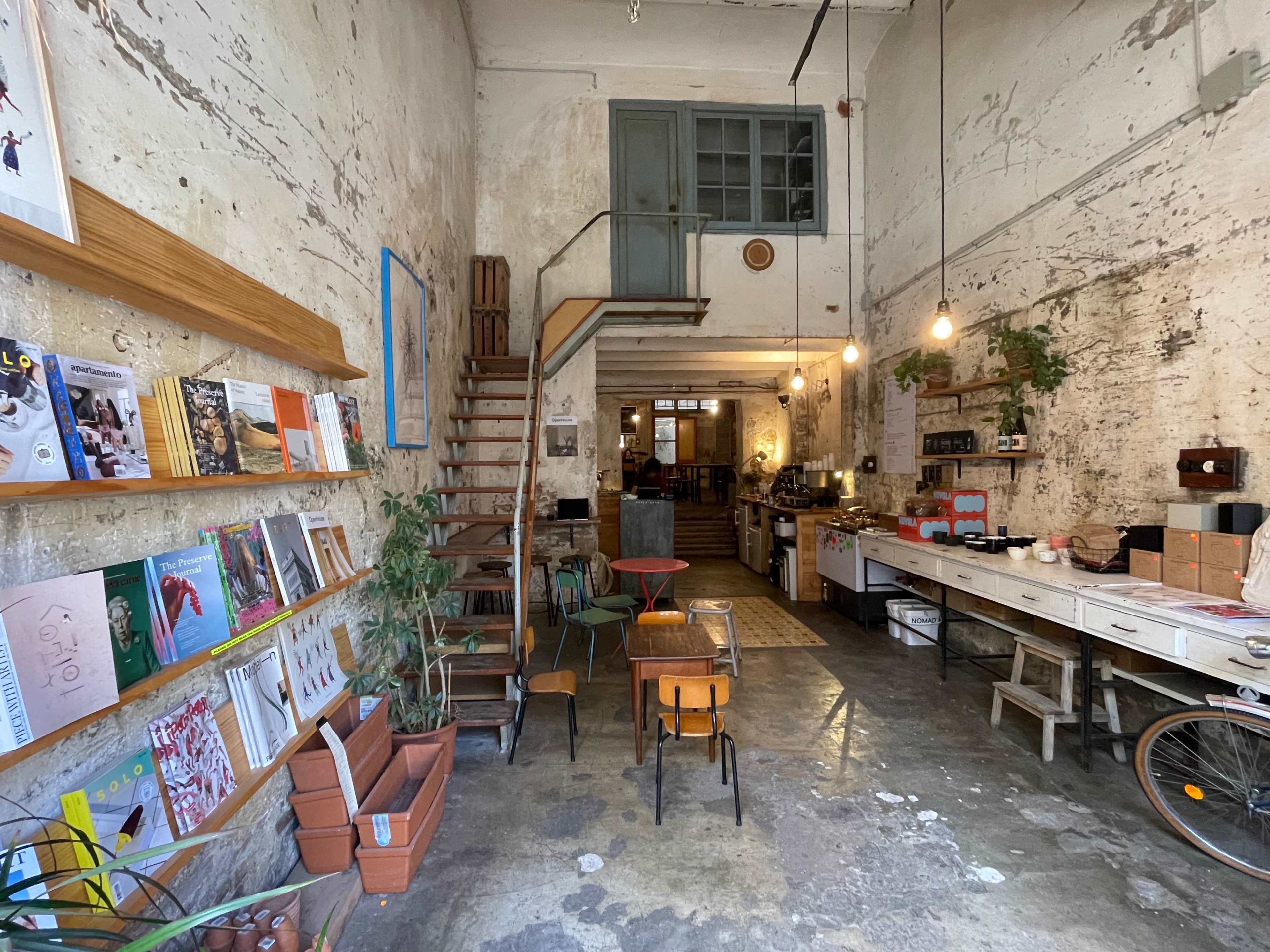 A rustic café with exposed brick walls, wooden stairs leading to an upper level, and shelves filled with books and magazines.