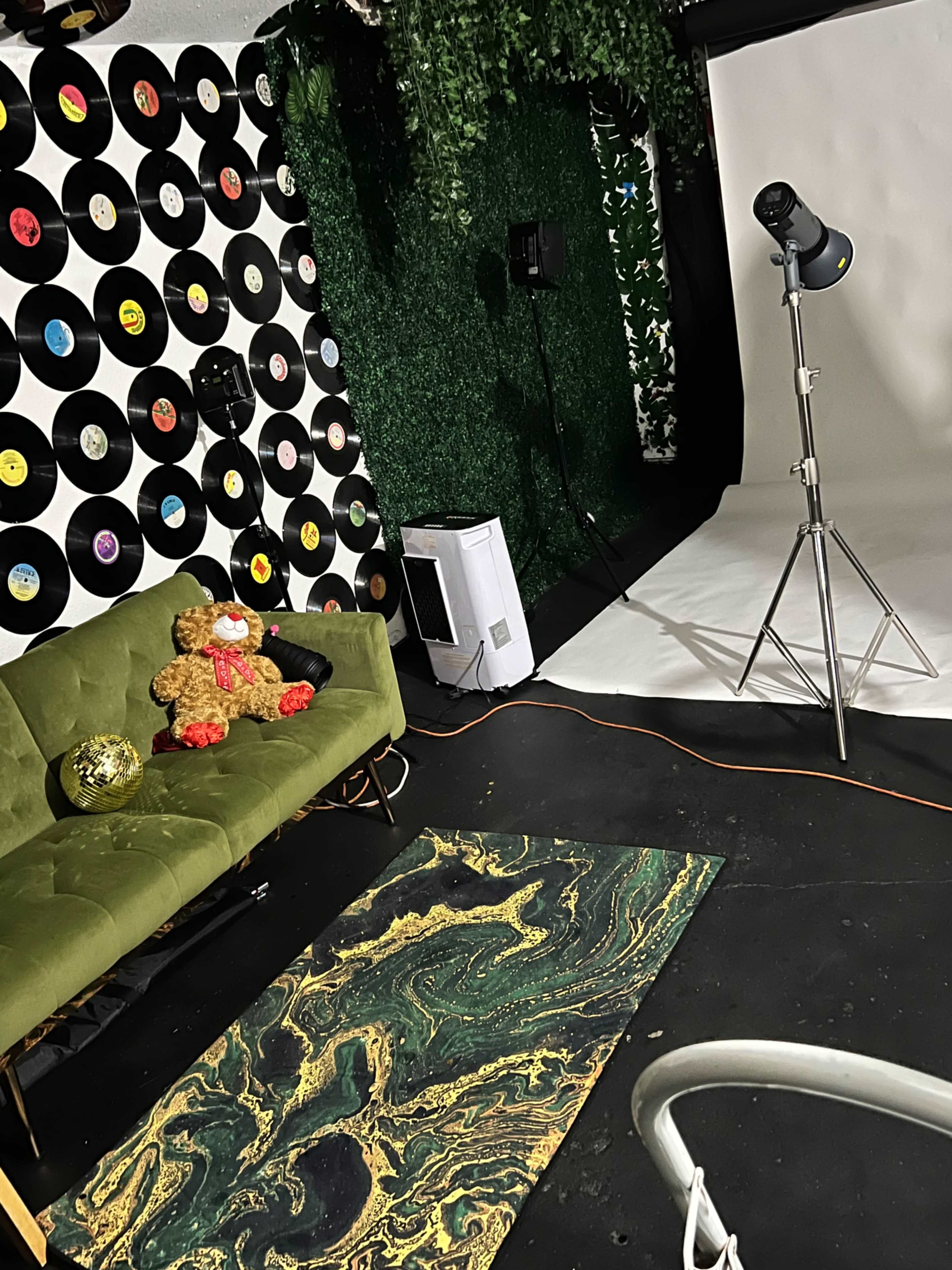 A photography studio with a green couch, a teddy bear sitting on it, a decorative rug, a backdrop of vinyl records on the wall, and a lighting setup.