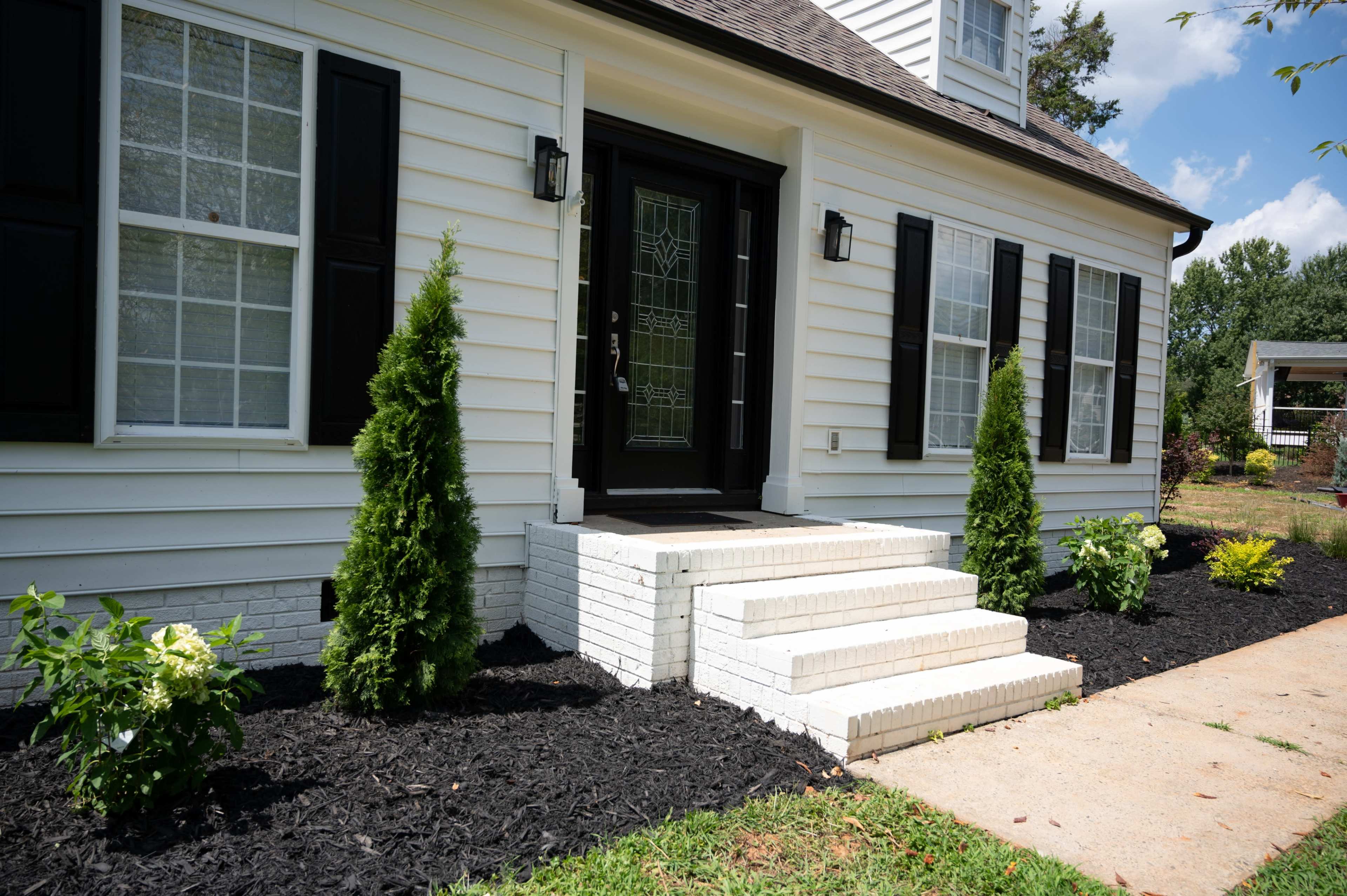 A white house with black shutters, featuring a front entrance with three steps and two small evergreen trees on either side of the doorway.