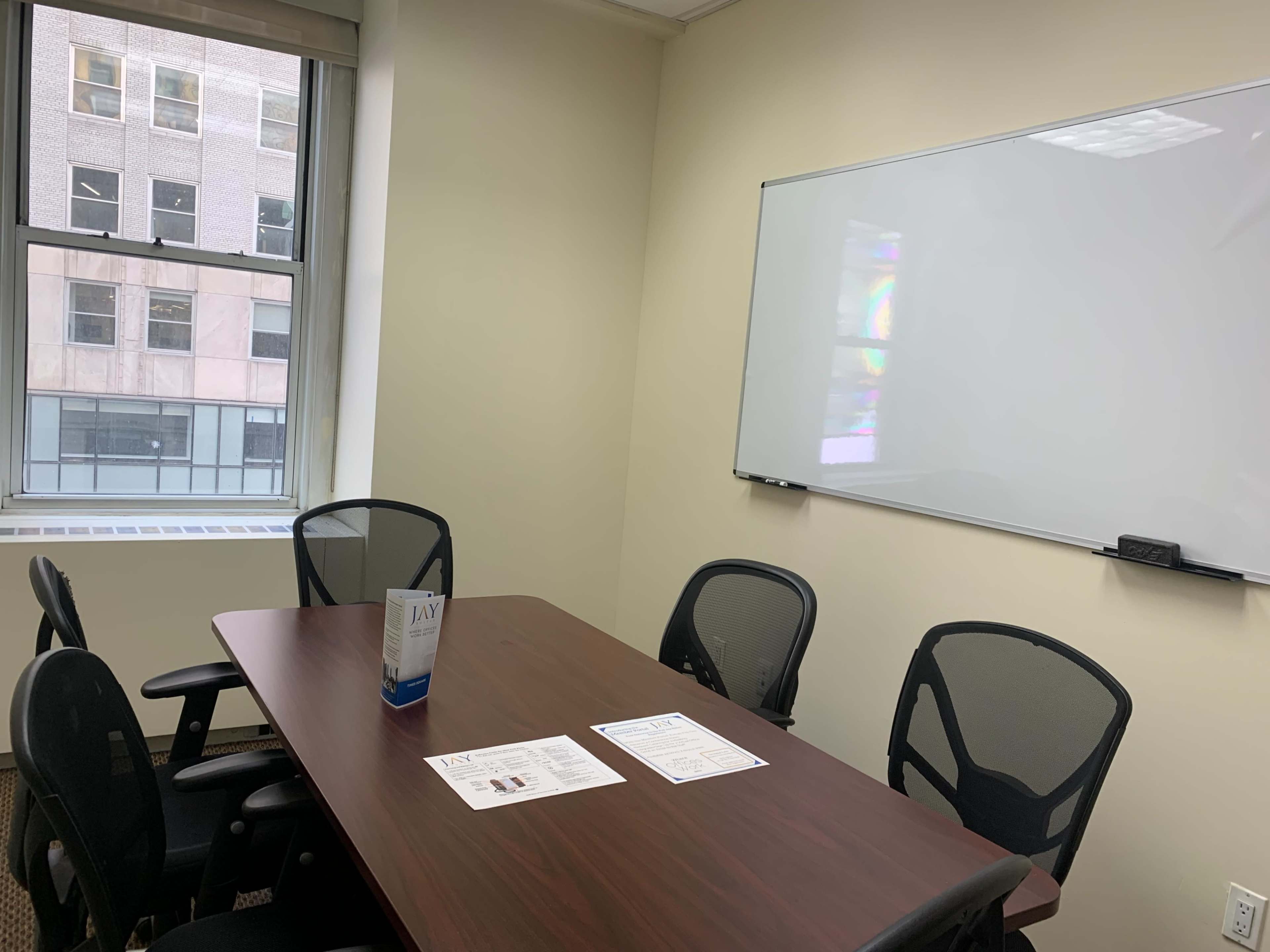 A small conference room with a wooden table, four black chairs, and a whiteboard on the wall, along with a few printed documents on the table.