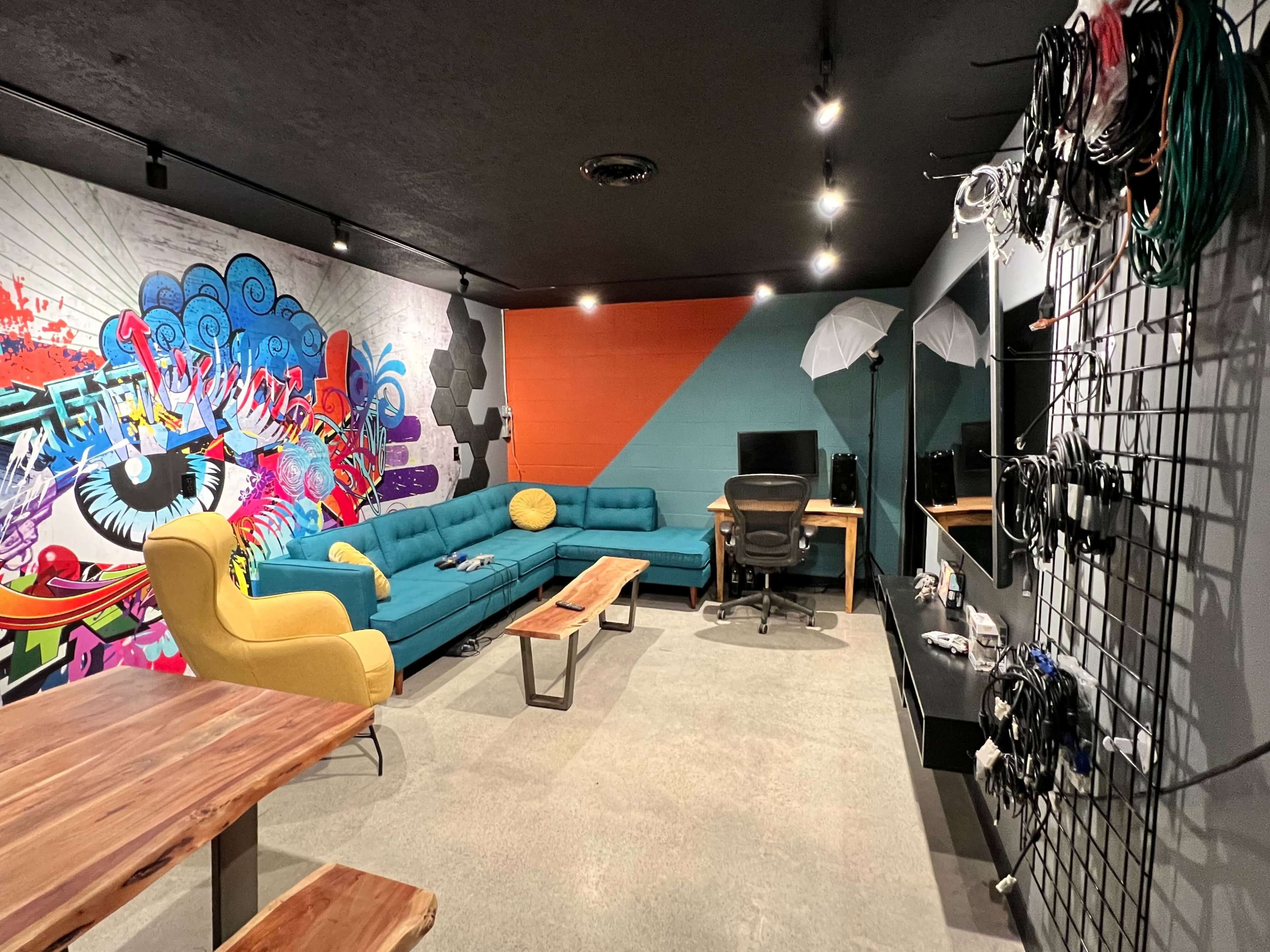 The image shows a modern creative workspace featuring colorful murals, a turquoise sofa, wooden tables, and organized equipment on the walls.