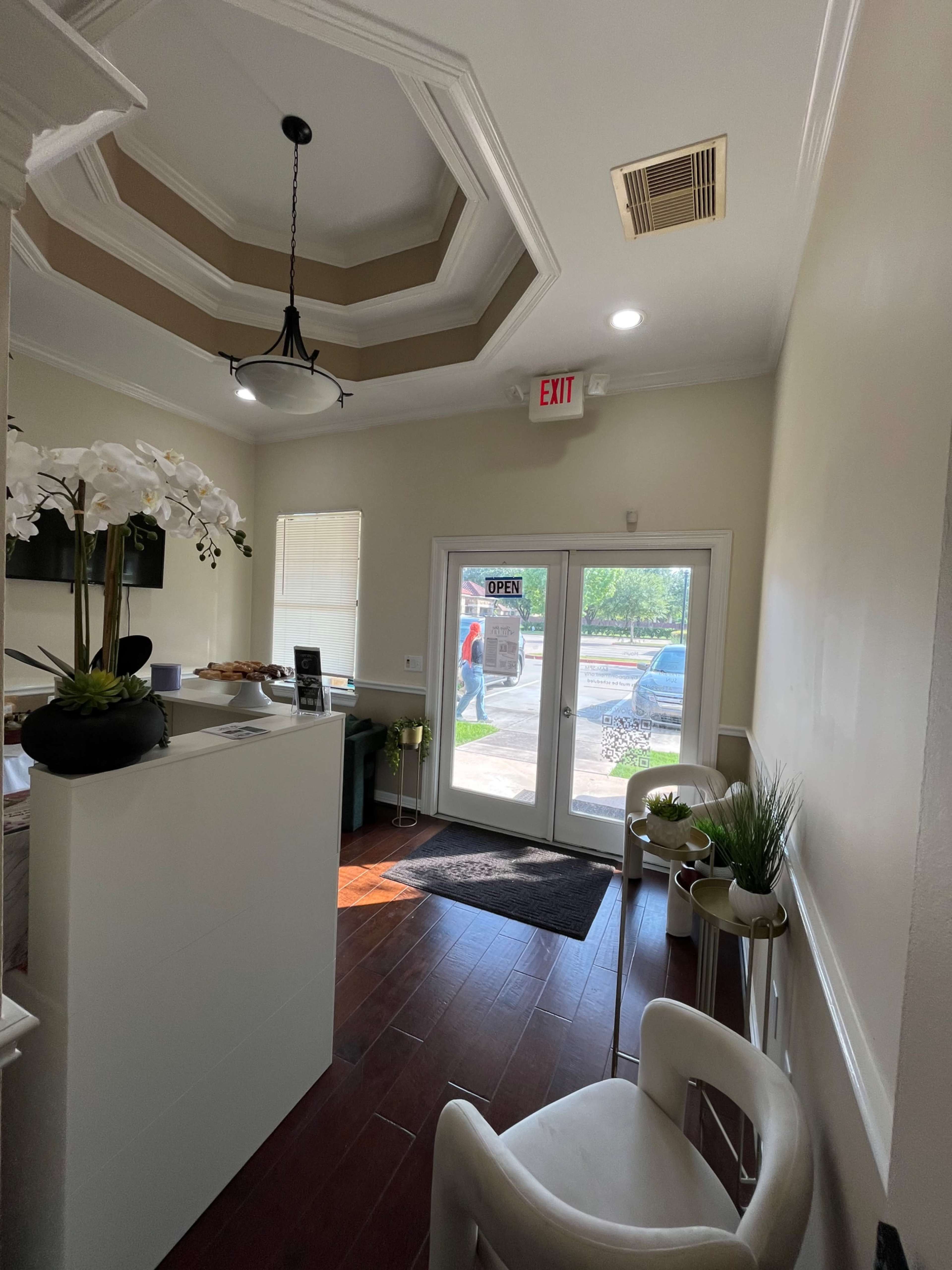 The image shows a well-lit reception area with a small desk, potted plants, and a large glass door labeled "OPEN."