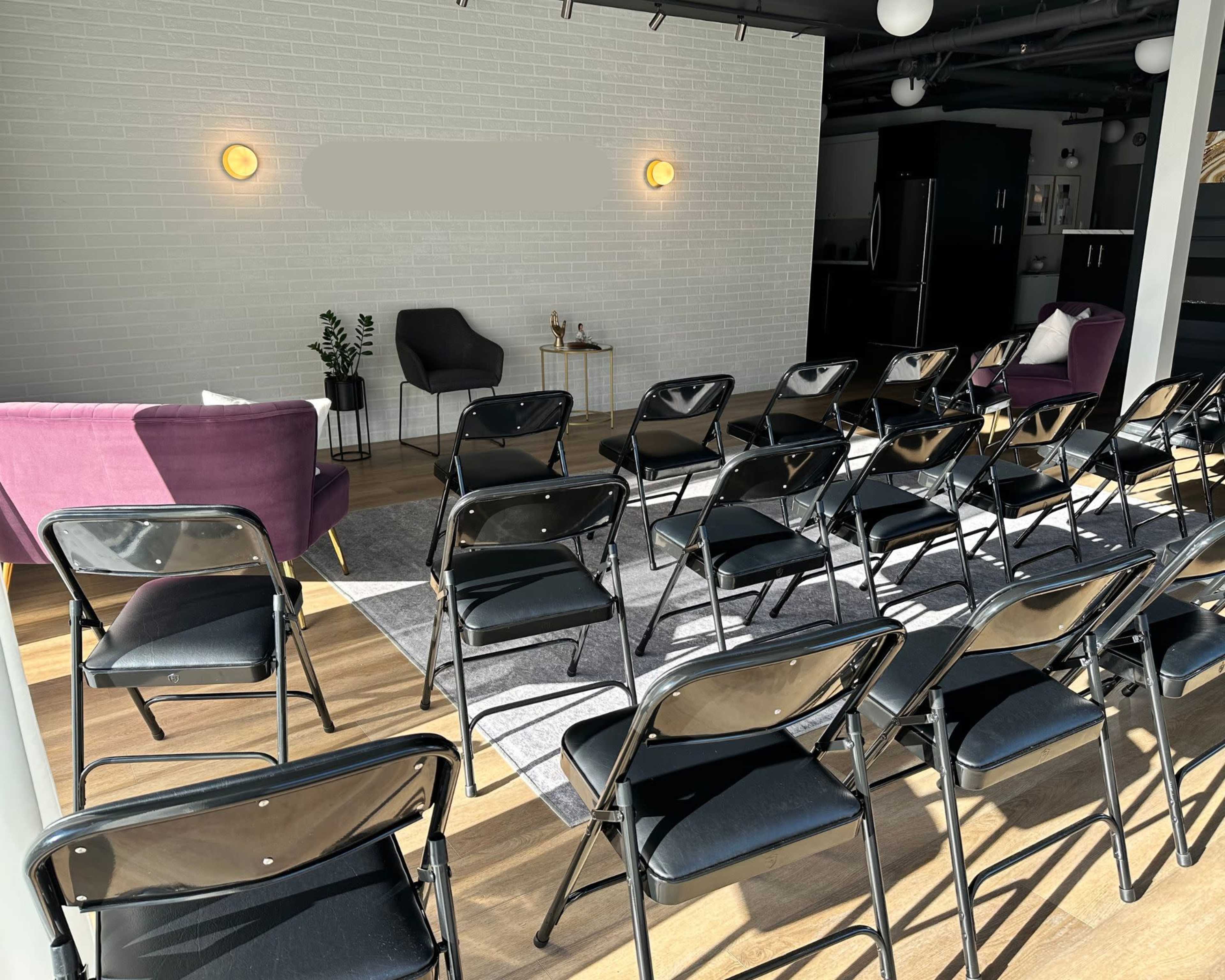 The image shows a neatly arranged seating area with several black folding chairs facing a small, minimalist stage set against a bright white wall, flanked by two purple armchairs.