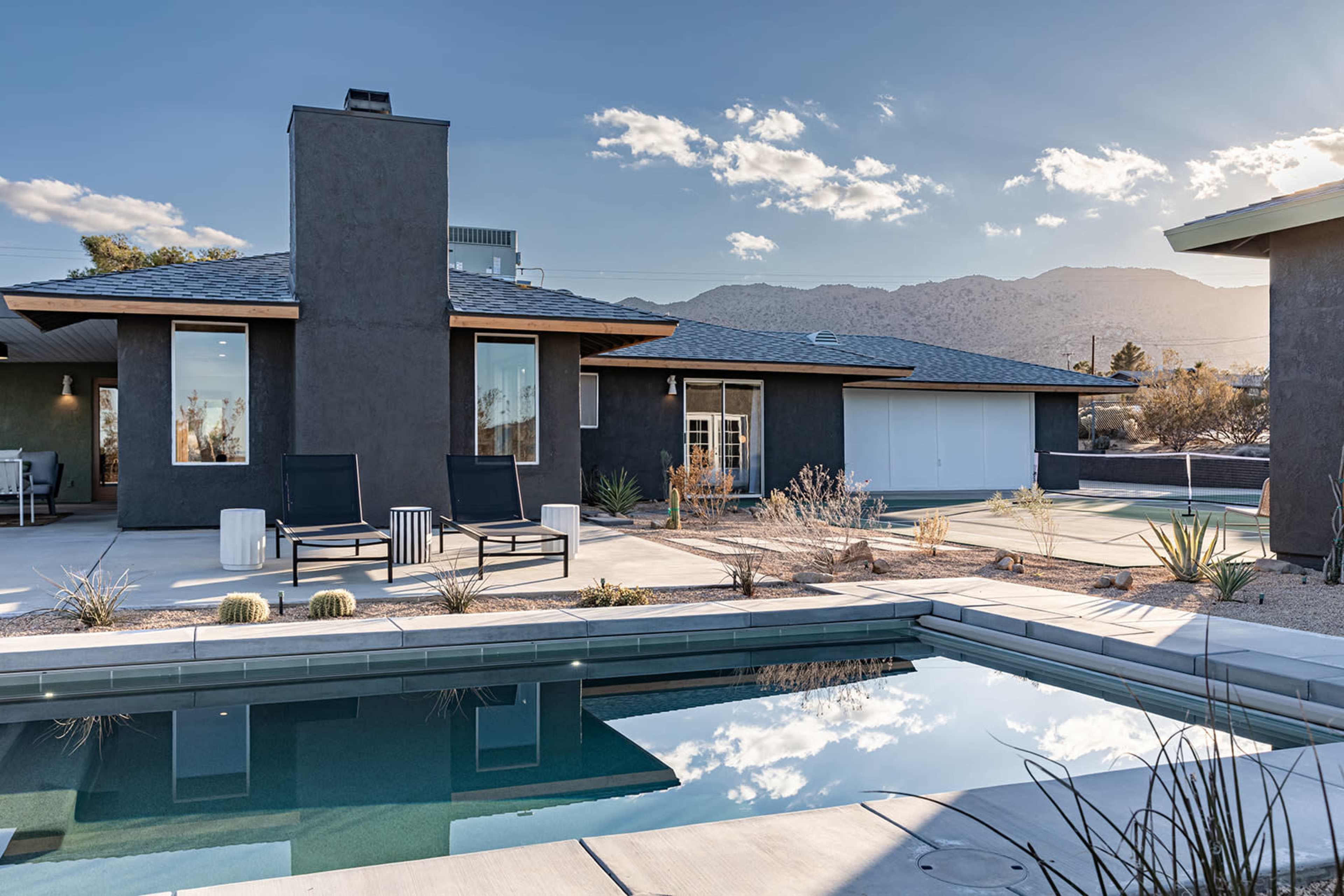A modern desert home with a swimming pool, surrounded by cacti and mountain views under a partly cloudy sky.