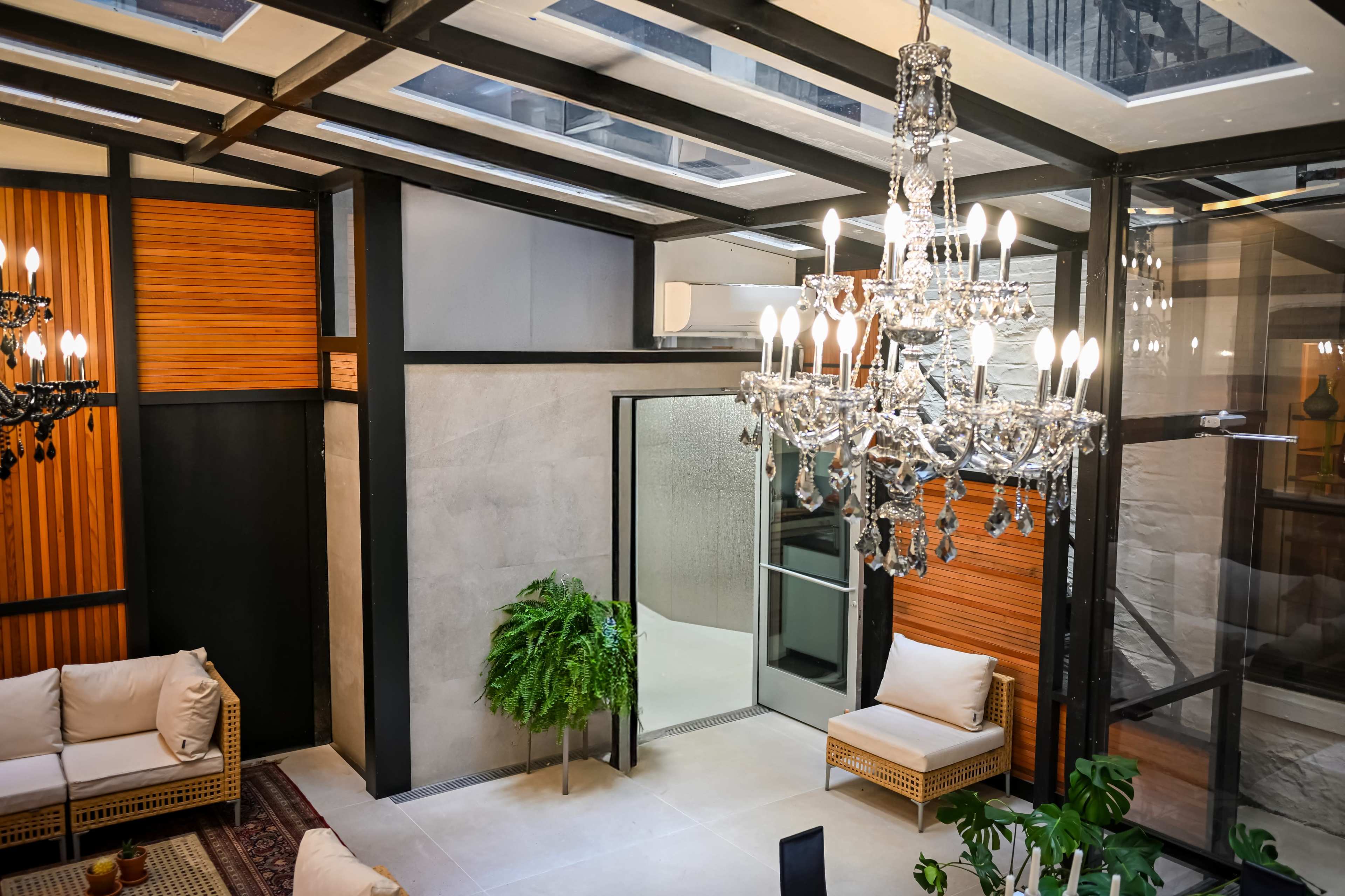 The image shows a modern indoor space featuring a glass ceiling, elegant chandeliers, and a blend of wooden and concrete materials with minimalist furniture.