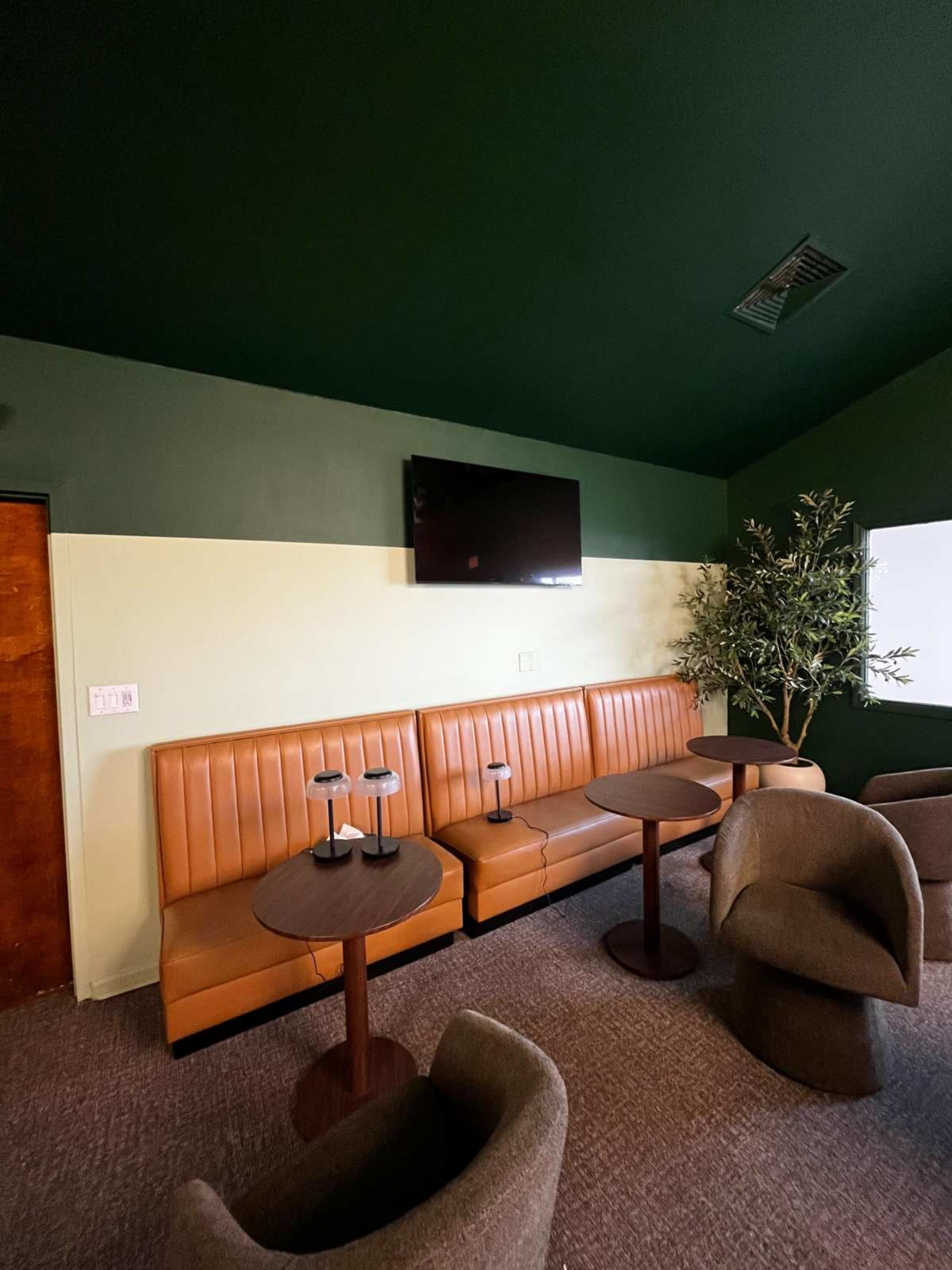 The image shows a cozy seating area with orange banquette seating and small round tables beneath a dark green ceiling.