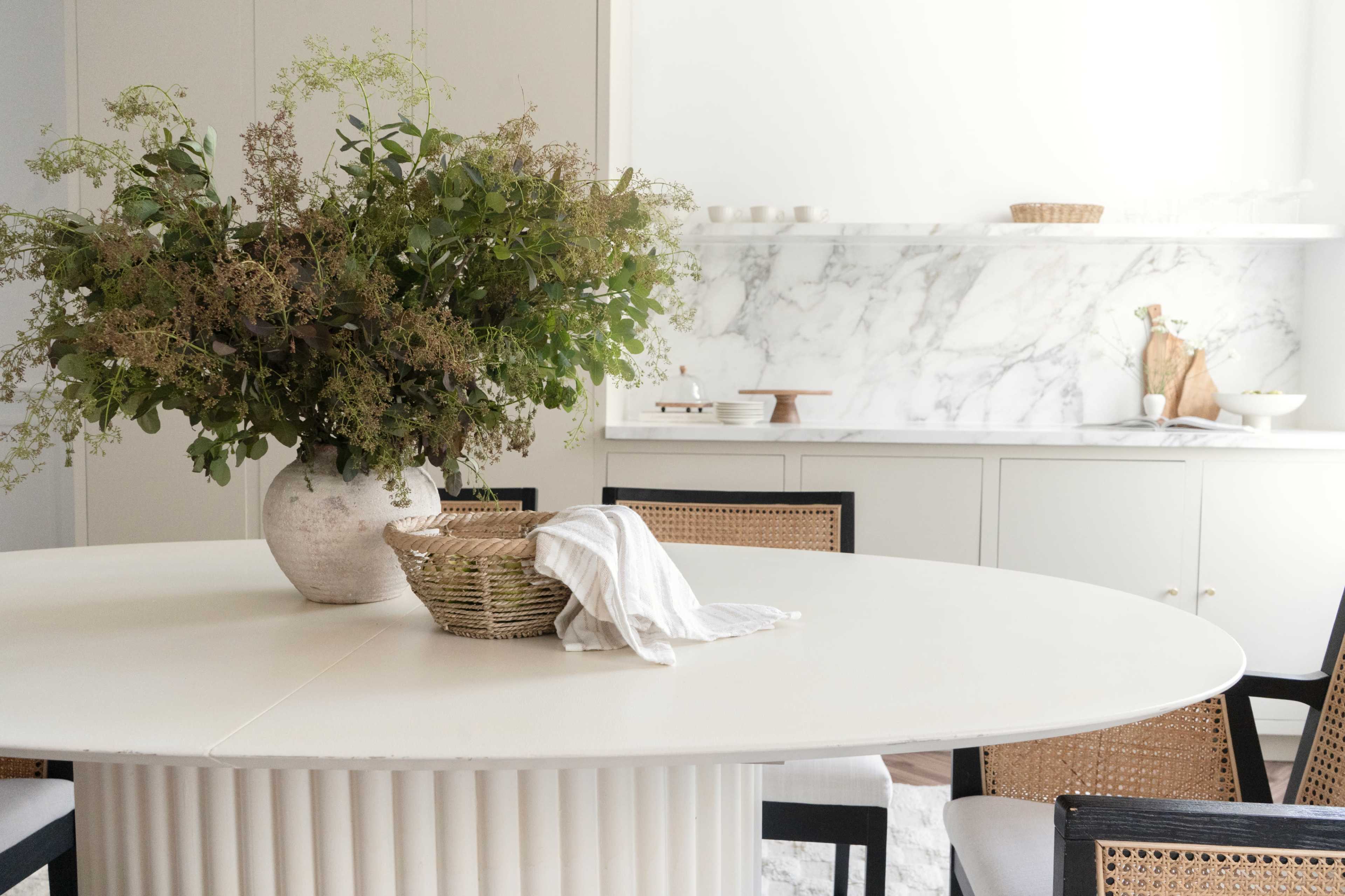 A large, round dining table with a white surface is adorned with a vase of greenery and a woven basket, surrounded by black chairs in a bright, minimalist kitchen.