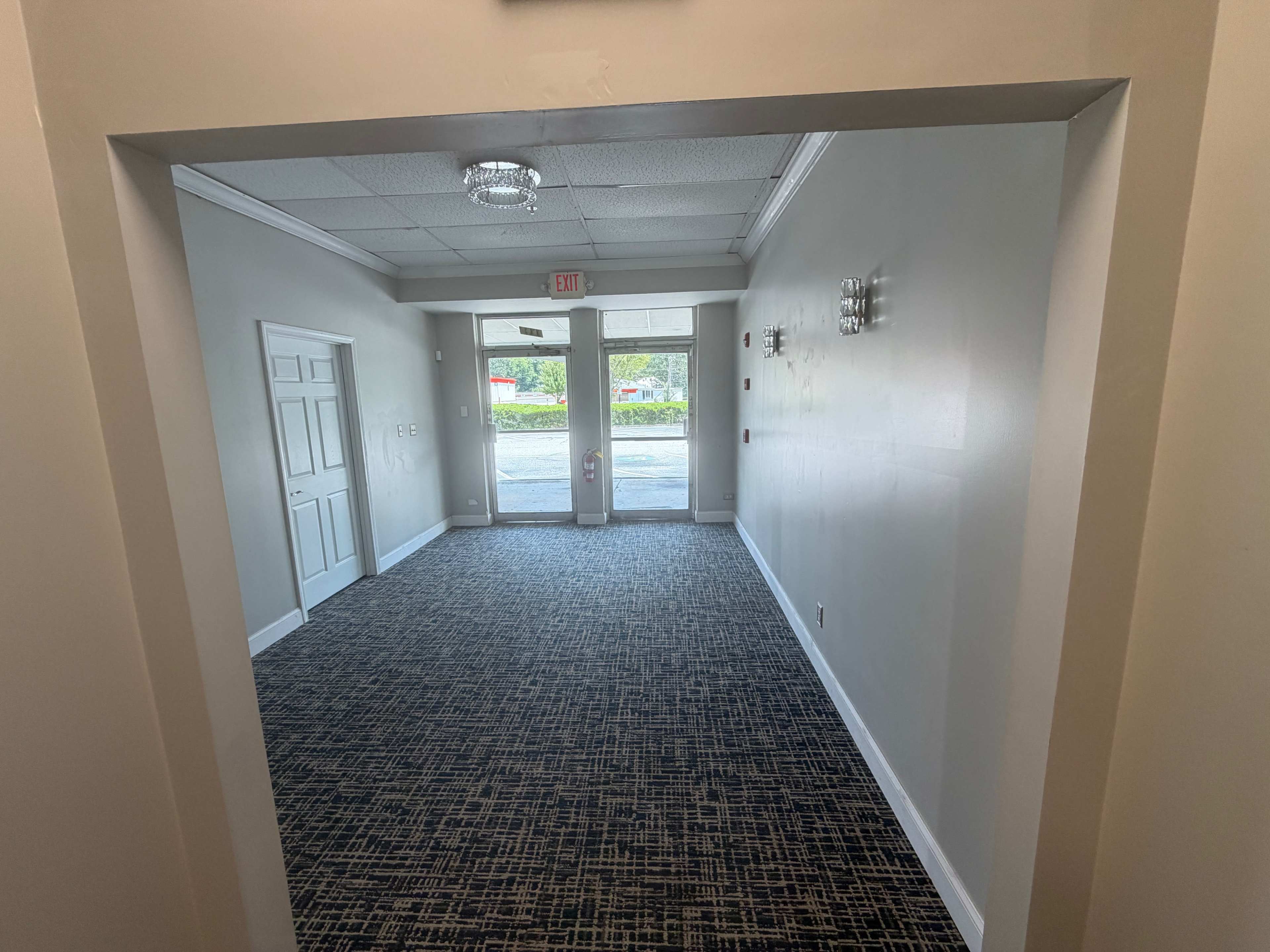 The image shows a view of a hallway with a carpeted floor, leading to a door with natural light coming from outside.