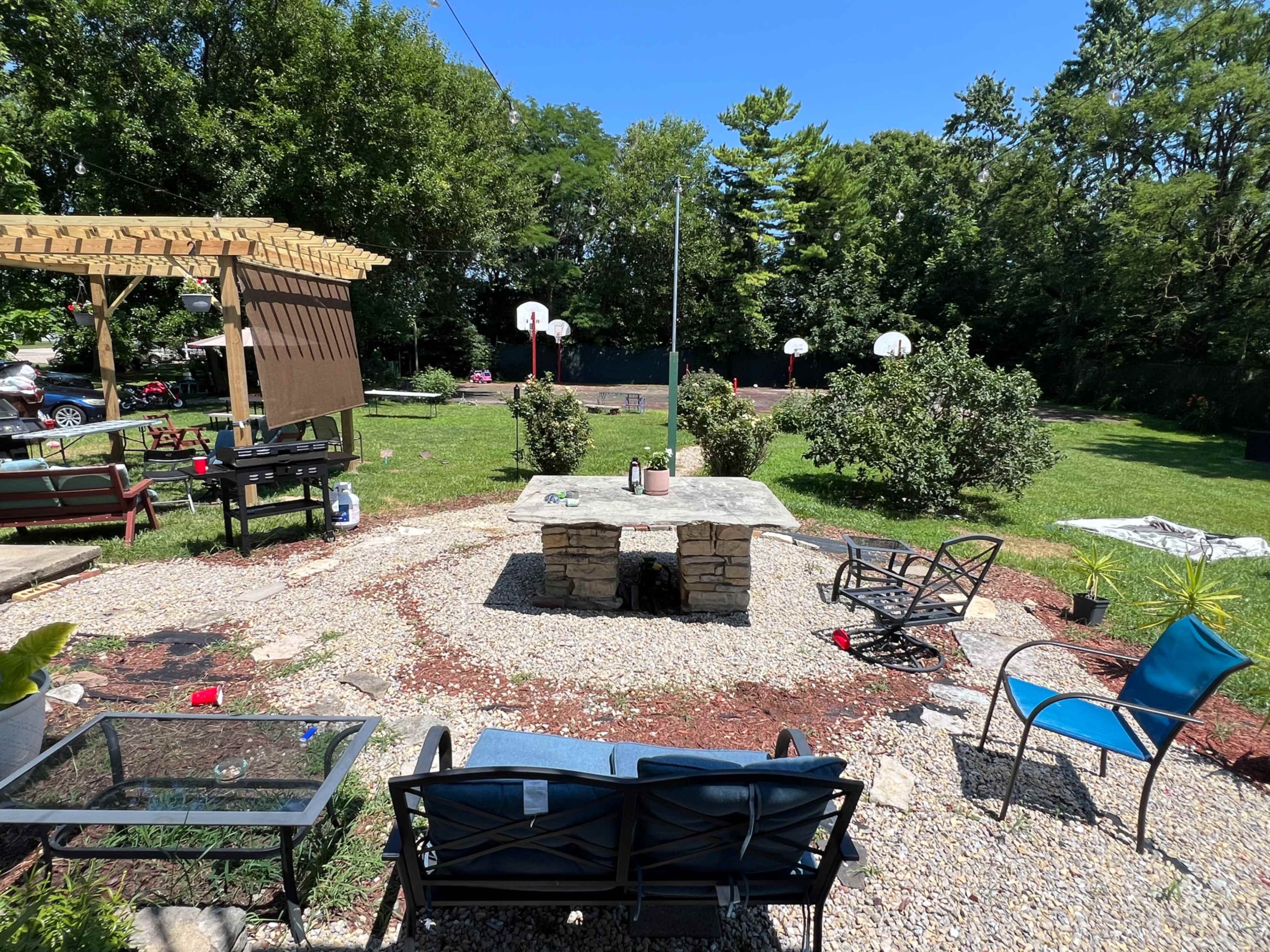 The image shows a backyard with a stone fire pit surrounded by seating areas, a grassy lawn, and basketball hoops in the background.