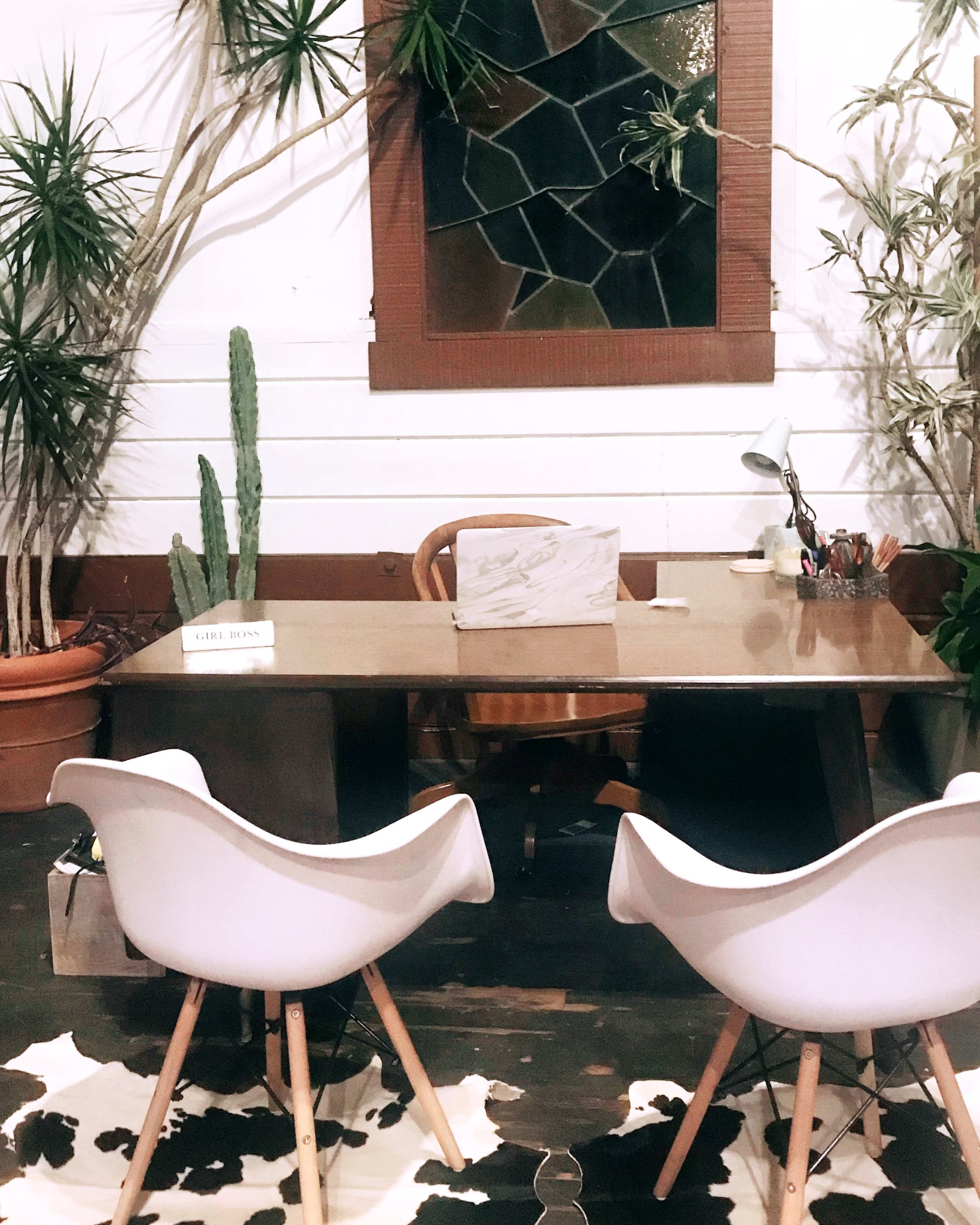 A minimalist office space featuring a wooden desk, two white chairs, and several potted plants against a wall with a stained glass window.