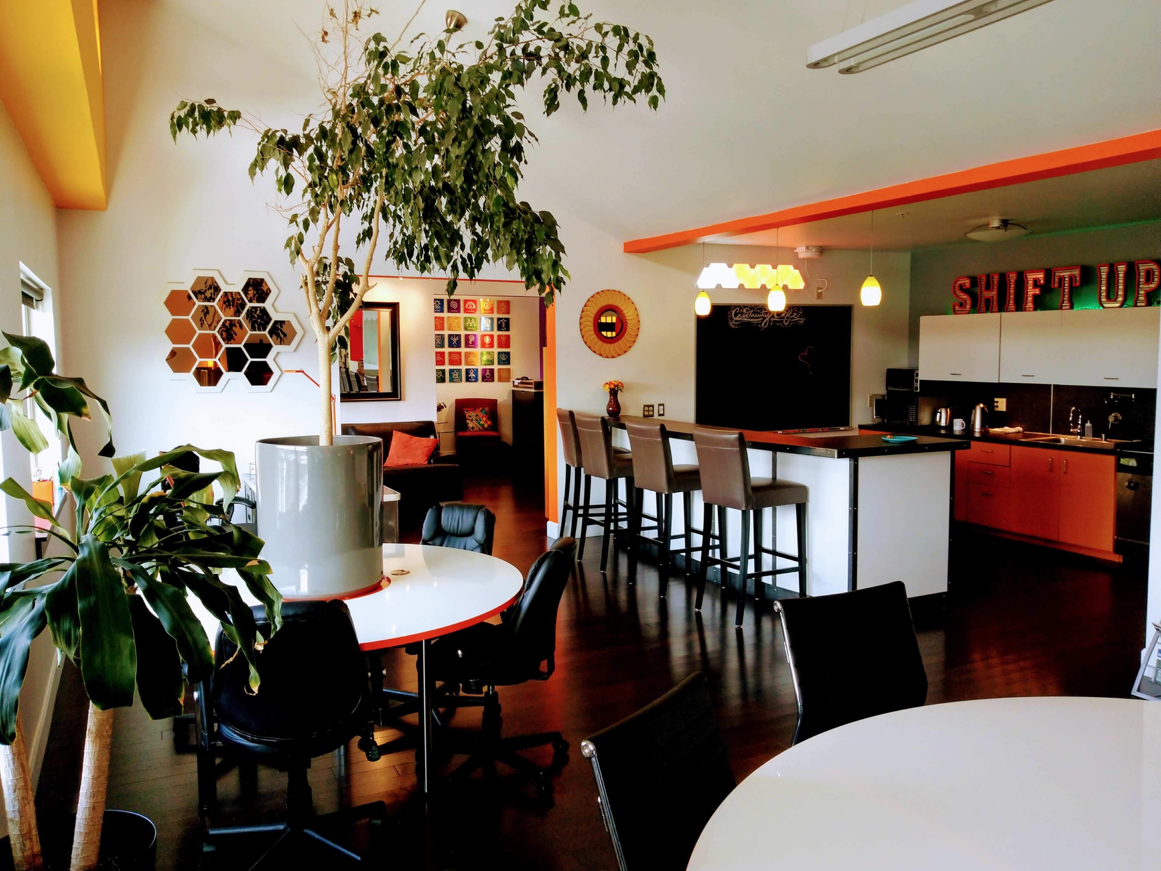 A modern open office space with a kitchen area featuring bar stools, a dining area with round tables, and decorative plants throughout.