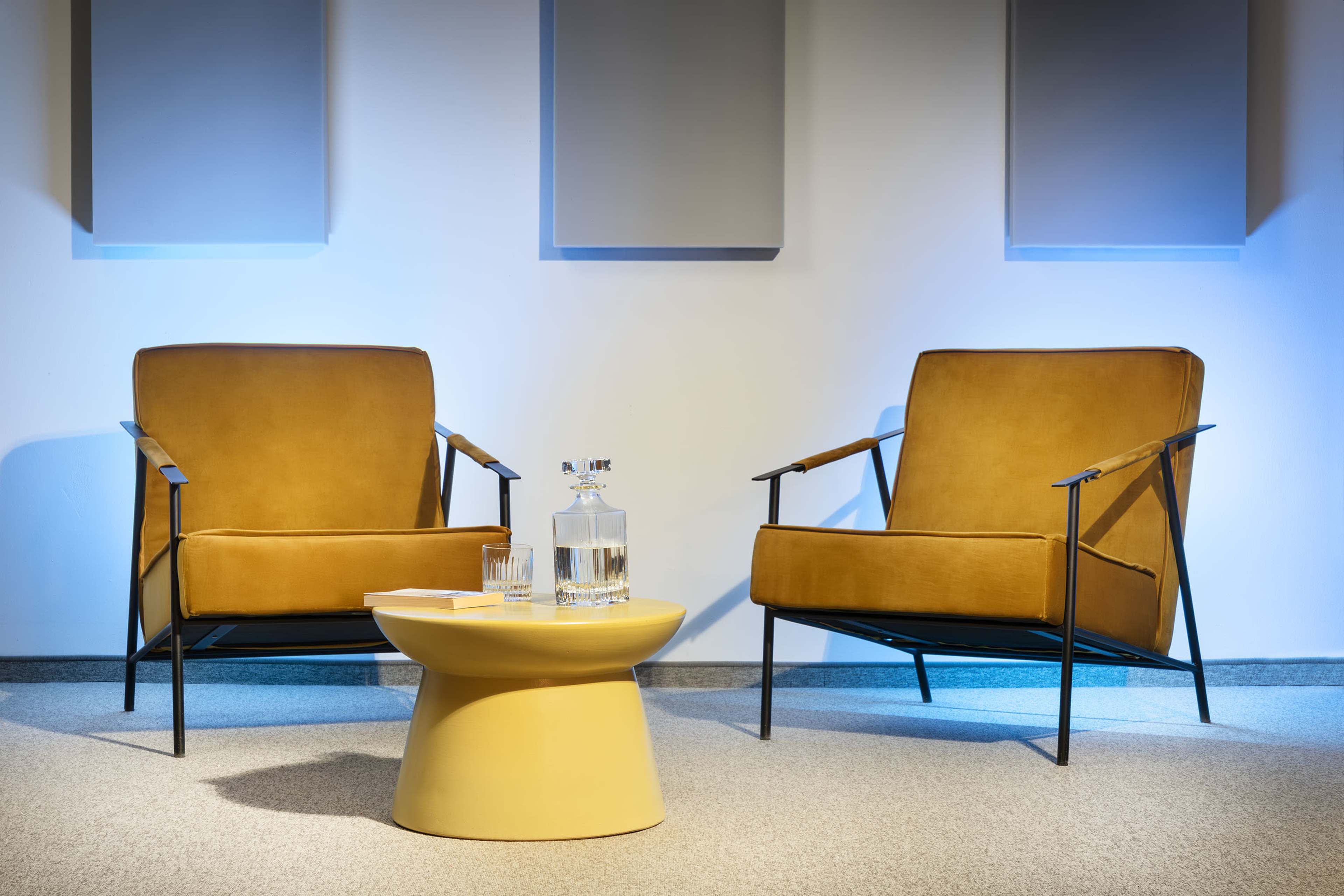 Two modern chairs with brown upholstery are positioned on either side of a round, yellow table, topped with a glass decanter and glasses, against a minimalist backdrop with blue lighting.