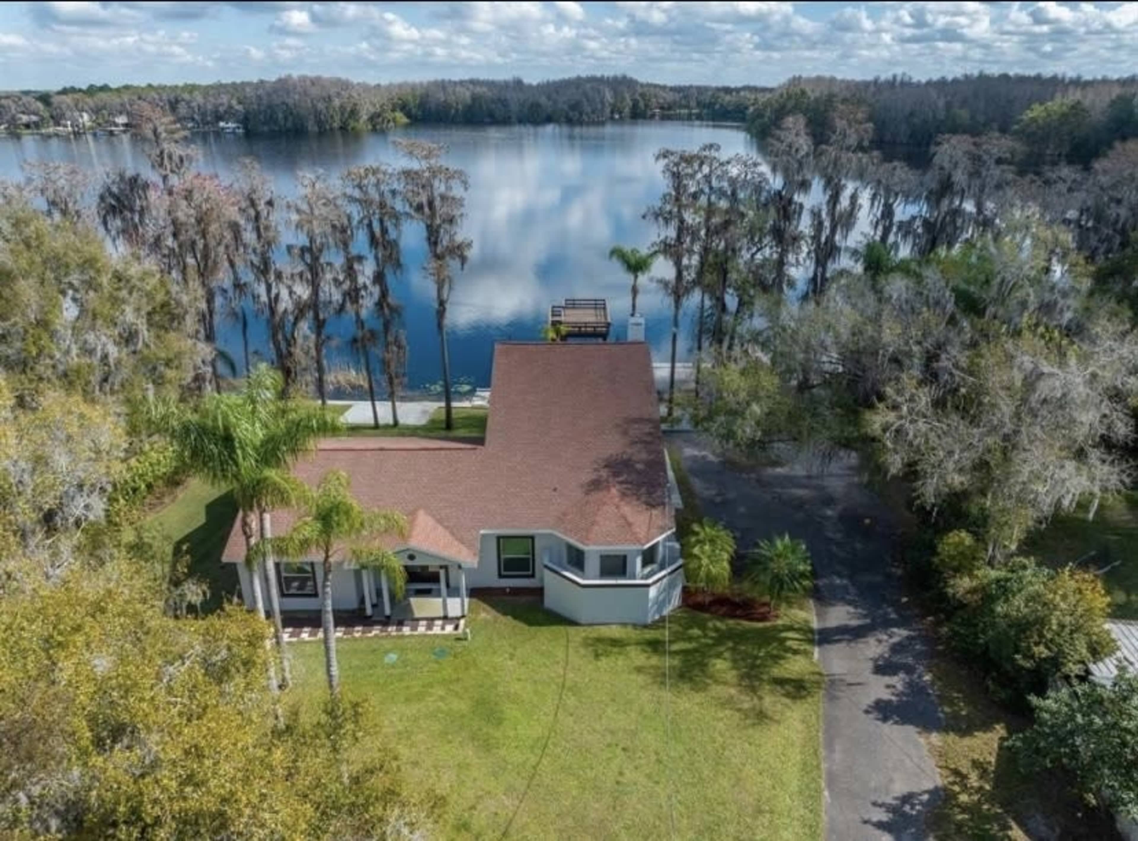 Aerial view of a single-story house with a red roof beside a calm lake surrounded by trees.